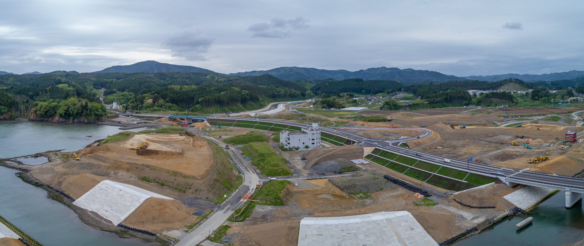 川の近くに広がる建設現場をパノラマで捉えた写真で、複数の道路や橋が建設中であり、重機も見られ、背景には丘や山々が広がり、土地が整地されインフラ整備が進む様子が写された風景の写真