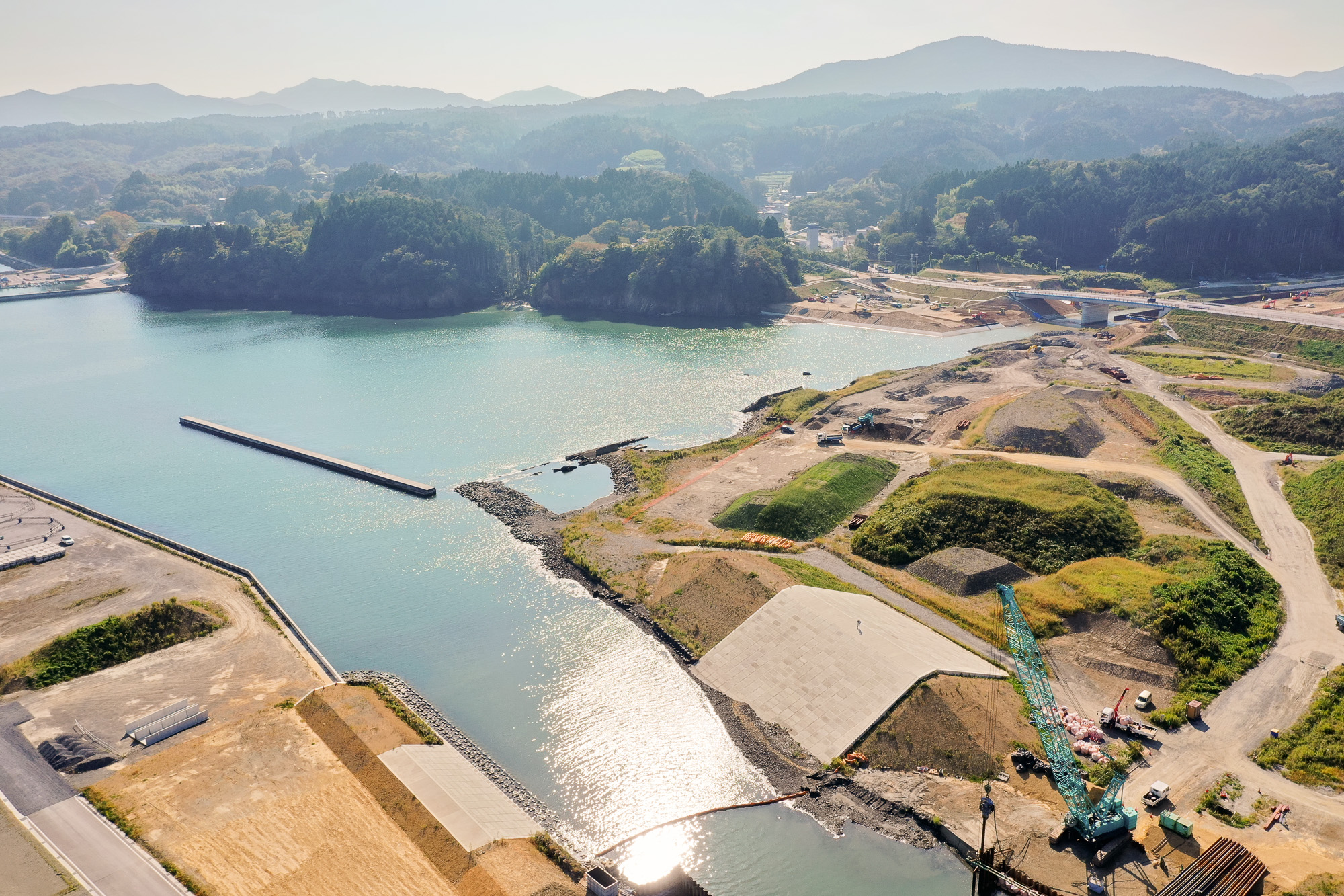 入り江と防波堤、整地された土地、そして奥に建設工事が進む丘や道路が広がる湾の奥の写真