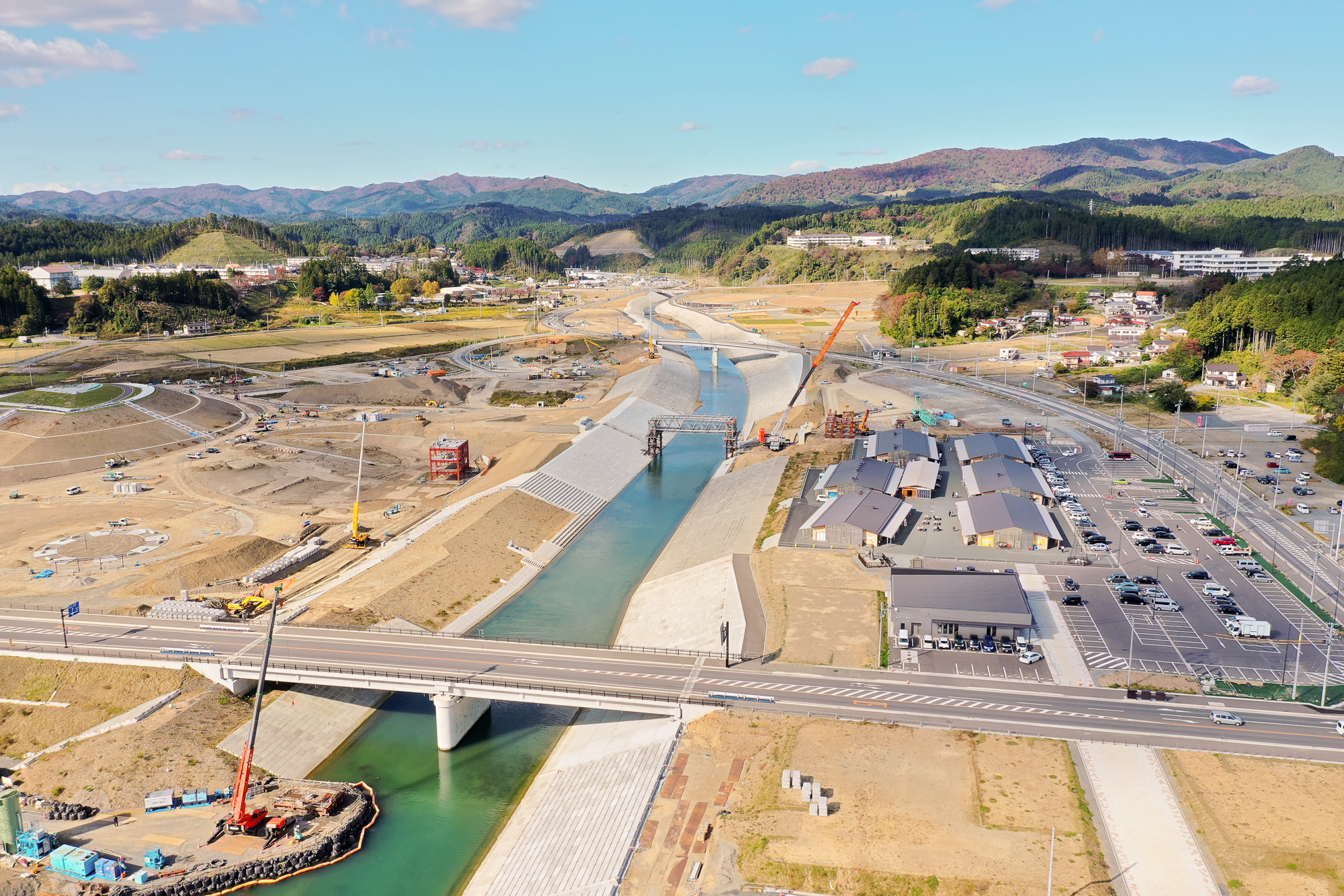 川の両岸がコンクリートで整備され、上流方向に向かって複数の重機が堤防工事を行い、右手には新しく建てられた木造風の商業施設と駐車場が並ぶ復興工事中の川沿いの写真