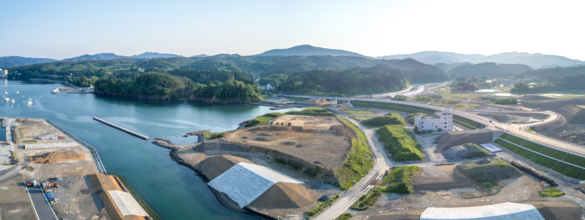 自然と人工構造物が混在する海沿いの地域を上空から撮影したもので、左側に水域と遠くに複数の船が見え、周囲には建設現場や道路、緑地が広がり、背景には木々に覆われた丘と山々、晴れ渡った空が広がる風景のパノラマ写真