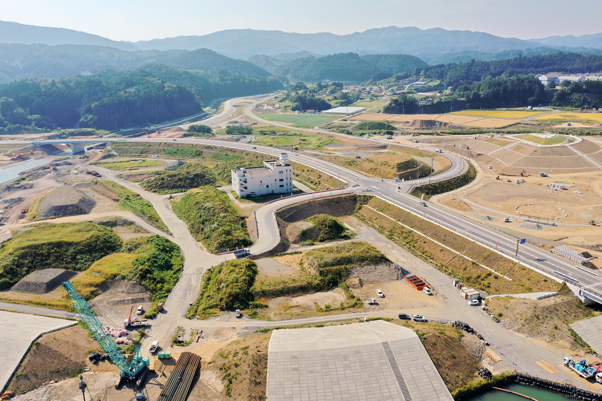 海に面した丘陵地帯に緑のクレーン車や工事資材があり、右奥には小さな川と橋が見える造成地の写真