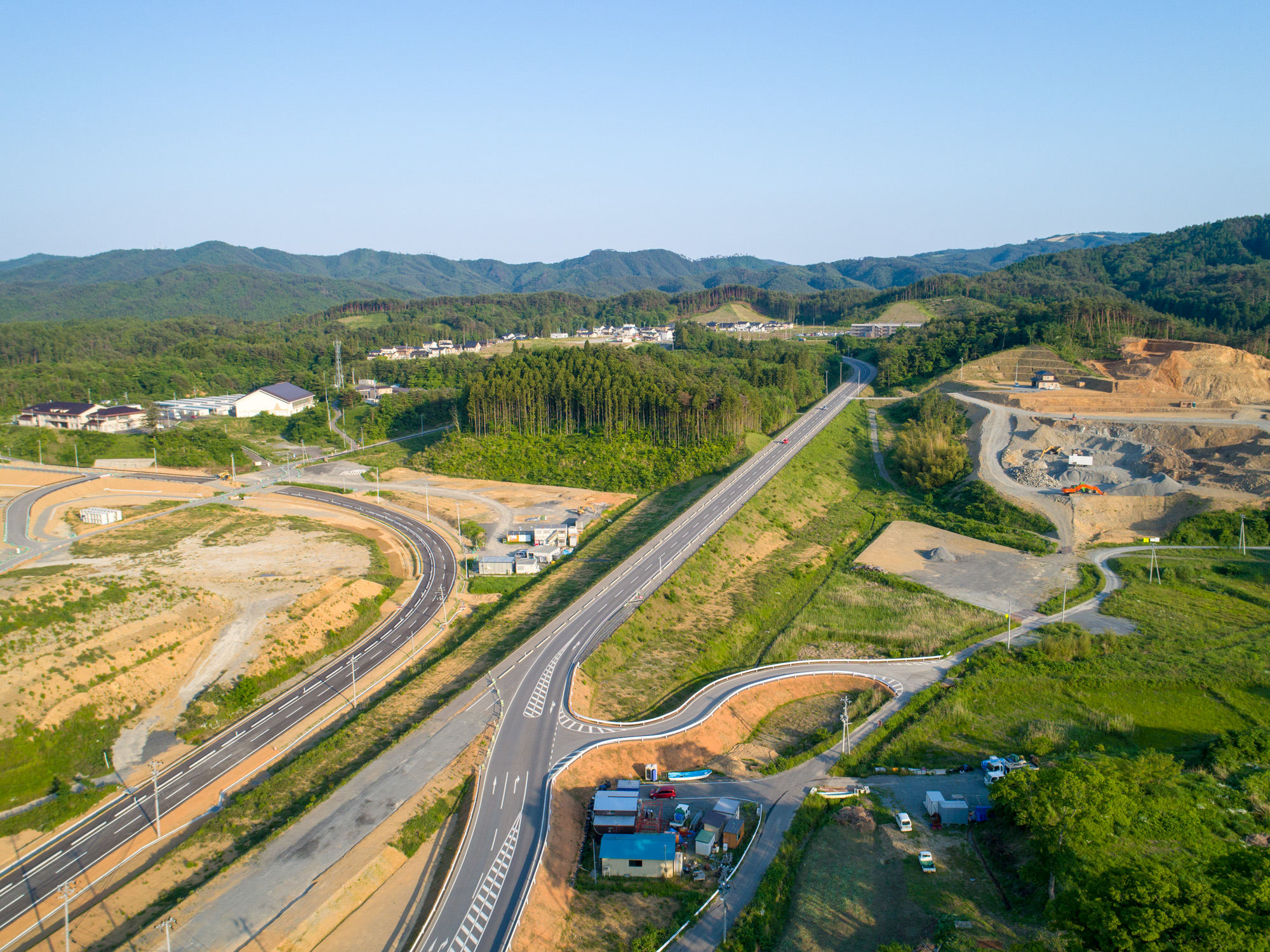 田園地帯を上空から撮影したもので、斜めに走る幹線道路と交差する曲がった道路、緑の畑や建設現場、点在する建物、背景には森林に覆われた丘と山々、青空が広がる風景の写真