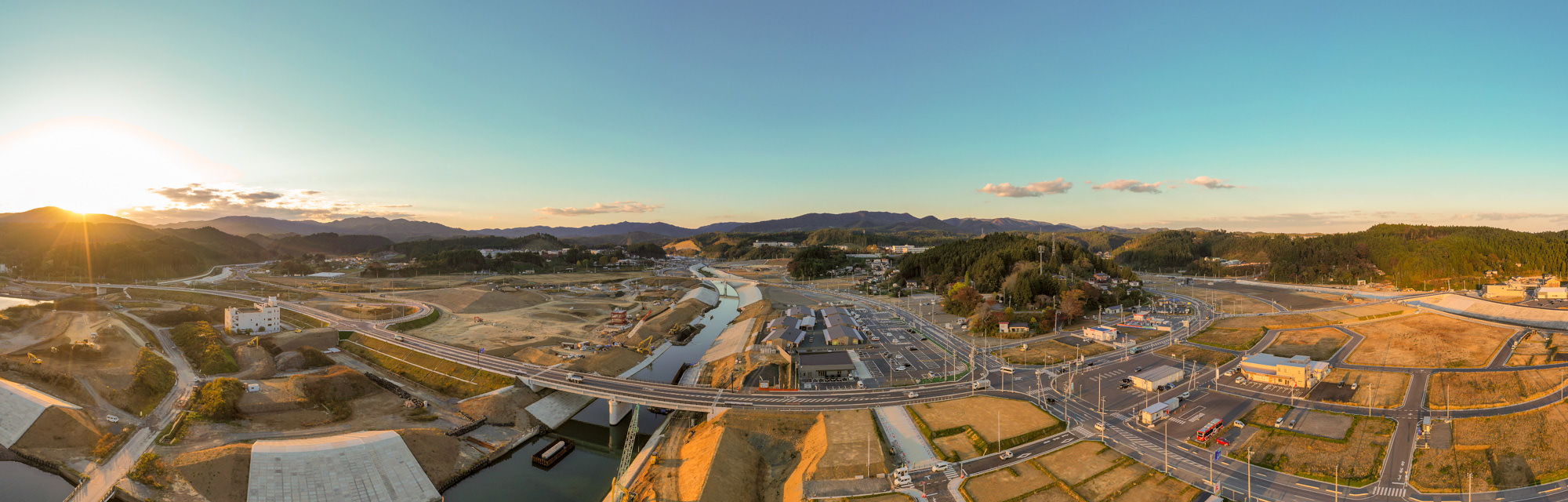 夕日に照らされながら、山々と新しい道路網、川、工事現場が広がる広大な地域の復興途中の町並みを上空から全体的に撮影した写真