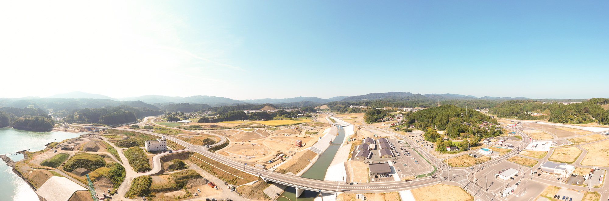 山に囲まれた沿岸部の町で、中央に川と整備中の土地が広がり、手前には海と道路が交差する様子が見える風景の写真