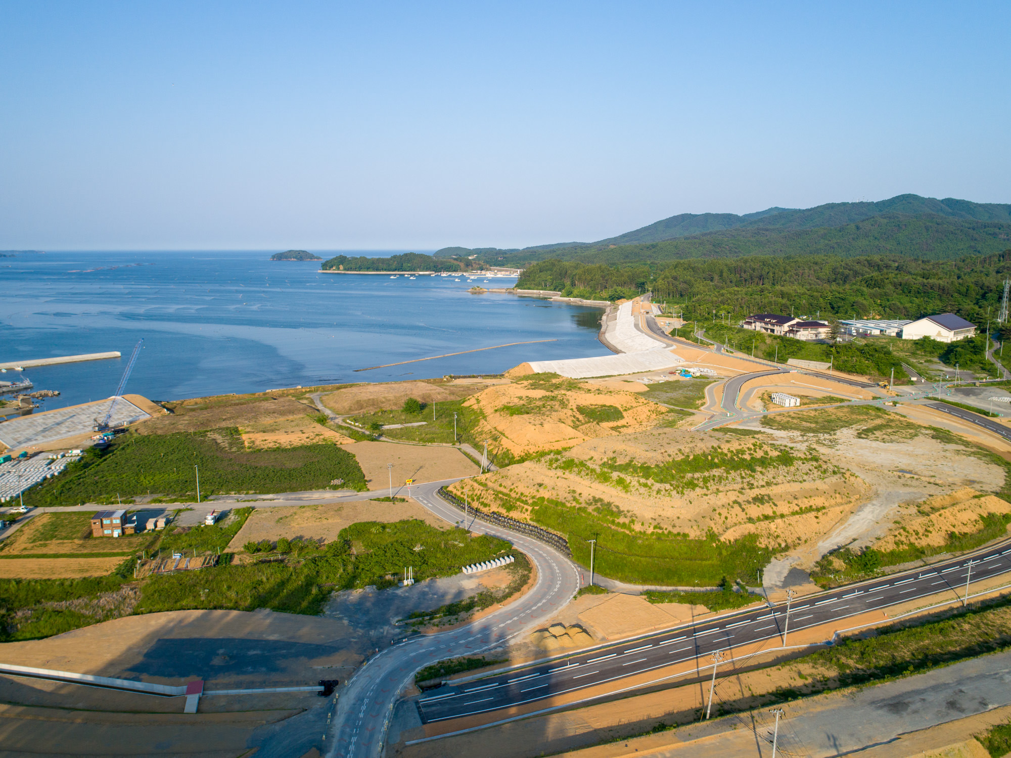 開発された土地と自然が混在する海岸地域で、道路や建物、緑地が点在し、遠くまで続く砂浜と木々に覆われた丘、穏やかな海と青空が広がる風景の写真
