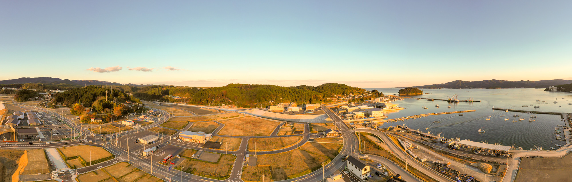 港に面した海岸沿いの町と海に浮かぶ漁船群、山や道路網、整備された造成地を含む町全体を夕方に上空から撮影した写真