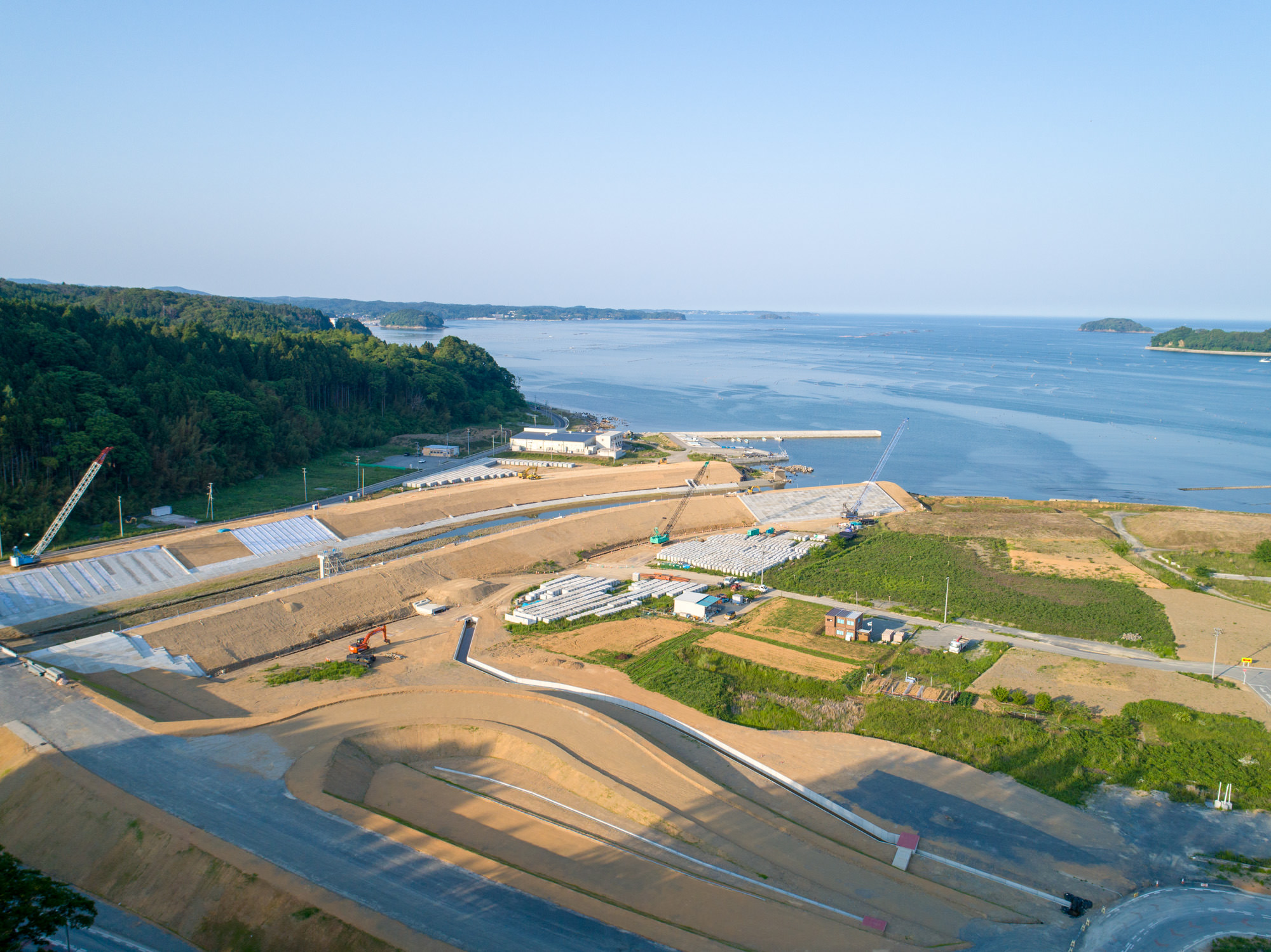 海岸沿いの工事現場を上空から撮影したもので、中央に建設機械や資材、左にクレーン、右に緑地と建物があり、背景には広がる海と遠くの島々、青空が広がる晴天の写真