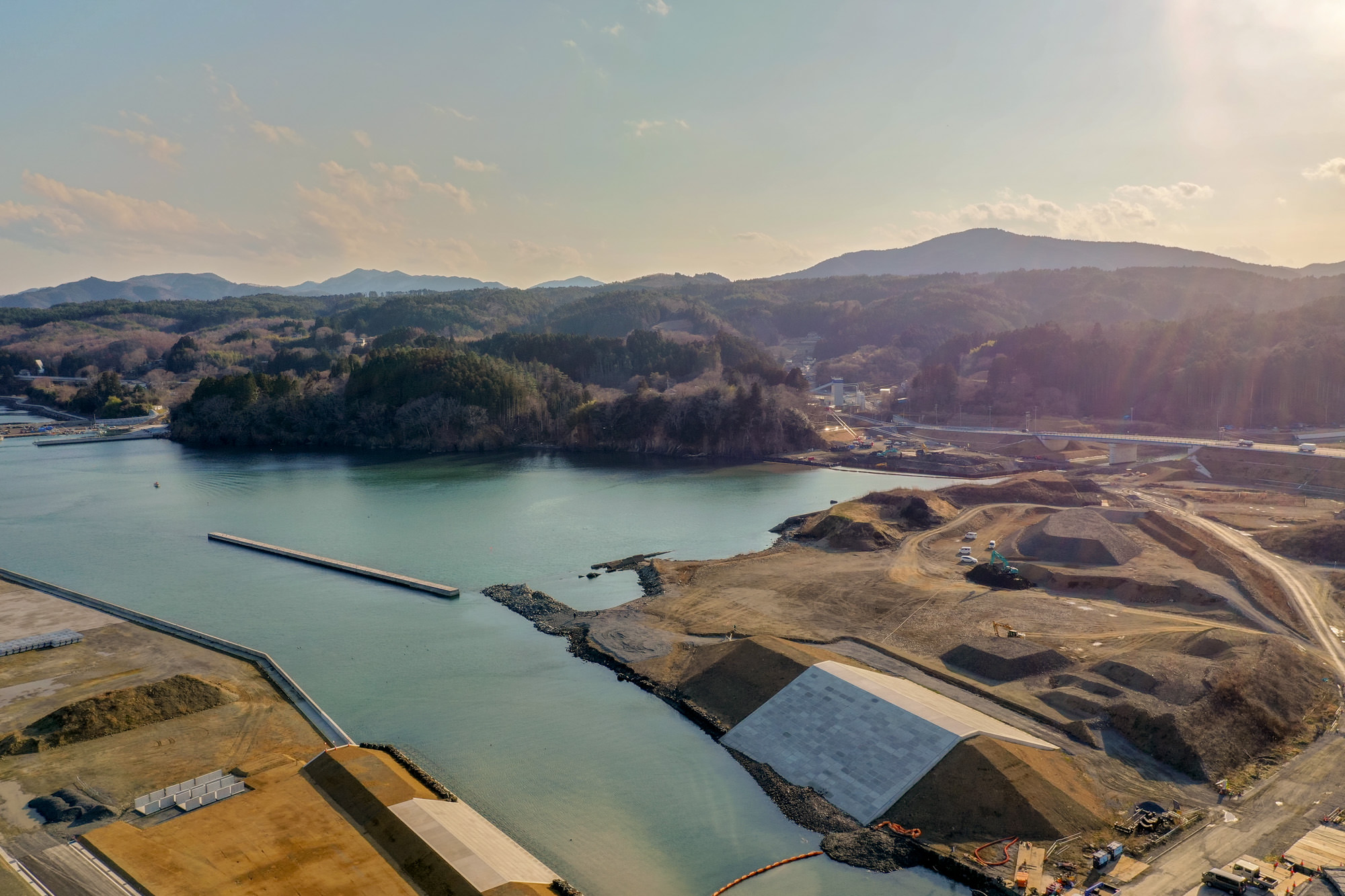 穏やかな水面の湾が陸地に囲まれて広がり、背景には丘や山が連なり、右側には工事中の様子が見られ、空には少し雲が浮かぶ湾の写真