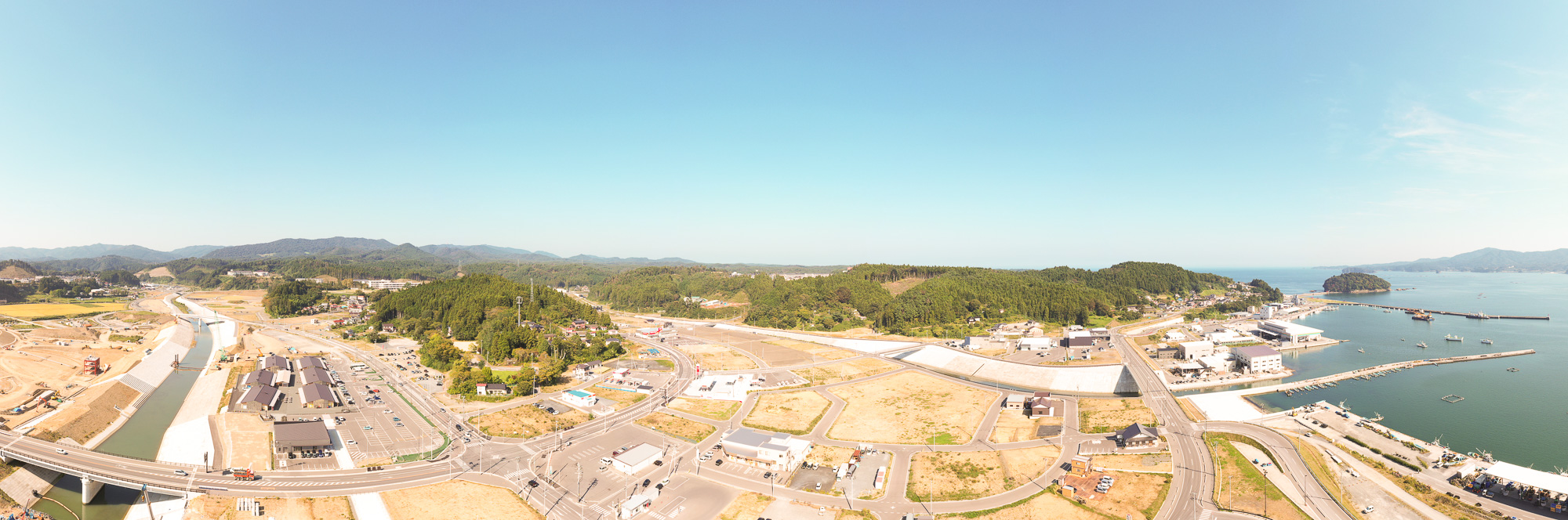 左に整備された川と造成地、右に海と港町が広がり、奥に山々が連なる様子を広くとらえた風景の写真