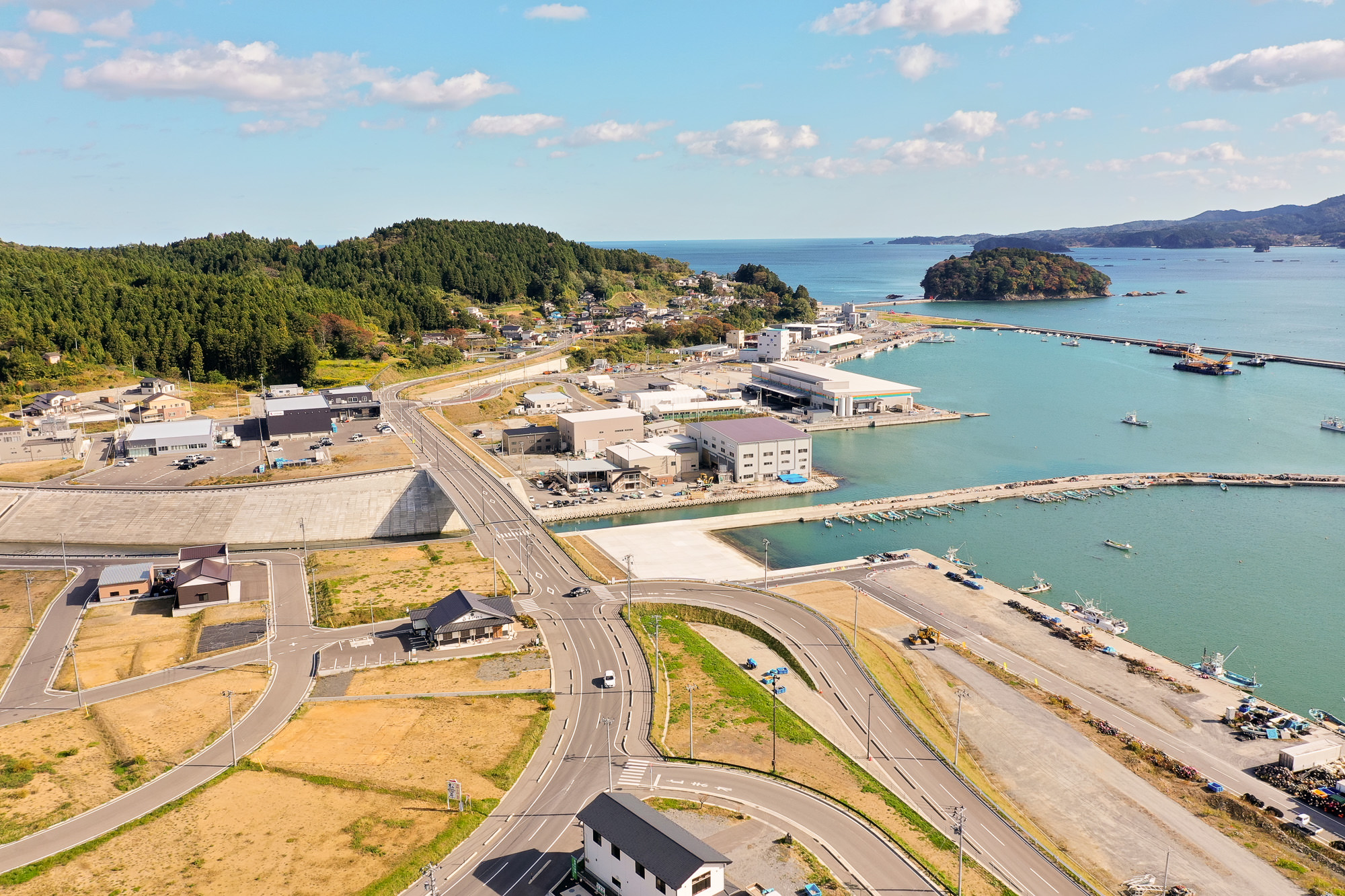 湾に面した港に複数の建物と倉庫が立ち並び、奥には小島が見え、前景には新しく整備された道路や住宅が並ぶ市街地沿岸部の写真
