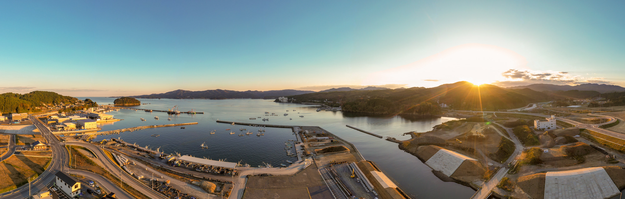 海に面した港町の夕方の風景で、静かな海に複数の船が停泊しており、背景には緑に覆われた山々と港町の建物が並んでいる様子が広がっている写真