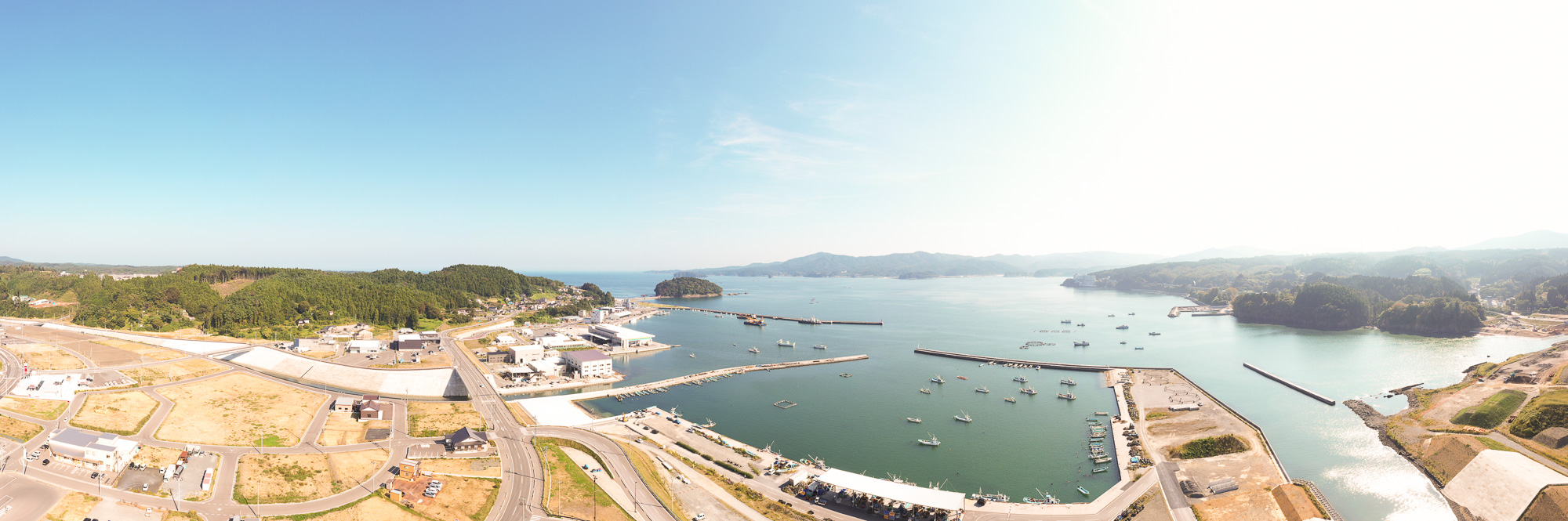 防潮堤と新しい道路が整備された港町の全景が海とともに広がり、右側には造成中の土地や橋が見える風景の写真