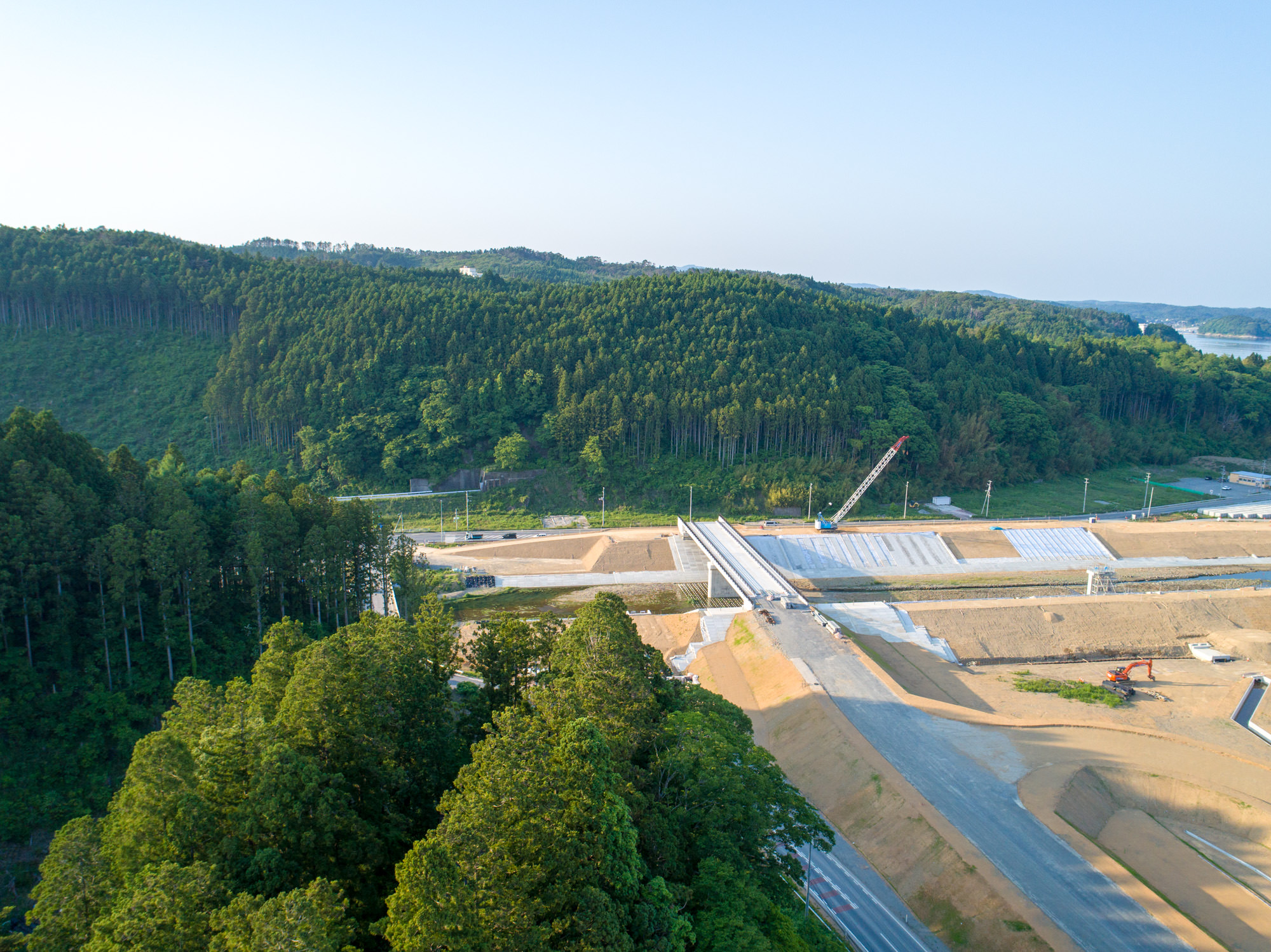 森林に囲まれた建設現場で、コンクリート構造物と土工が進む途中の橋があり、右側にはクレーンが見え、背景には青空の下に木々に覆われた丘が広がる風景の写真