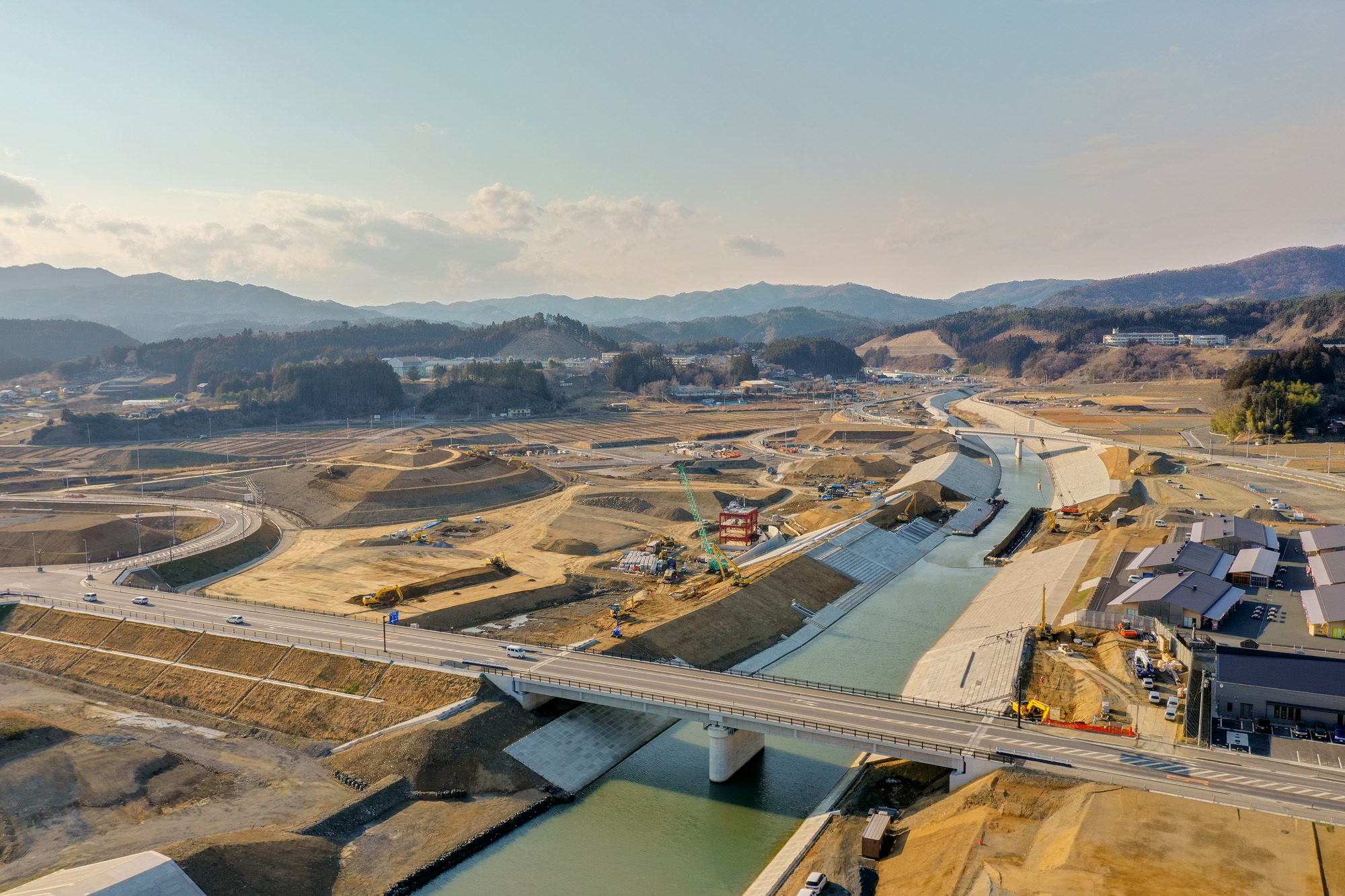 川に架かる橋を中心に、周囲に建設機材や資材、道路や建物が広がり、背景には山々が連なる起伏のある地形を上空から捉えた橋梁工事の写真