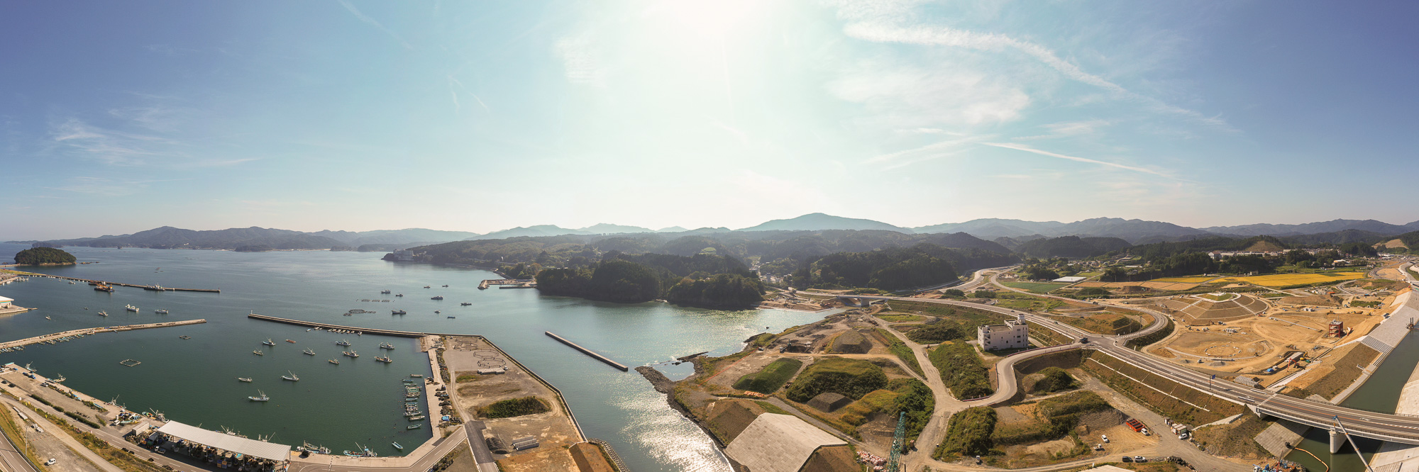 左手に漁港と漁船群、右手に山に囲まれた入り江と橋が見え、山並みが遠くまで続く風景の写真