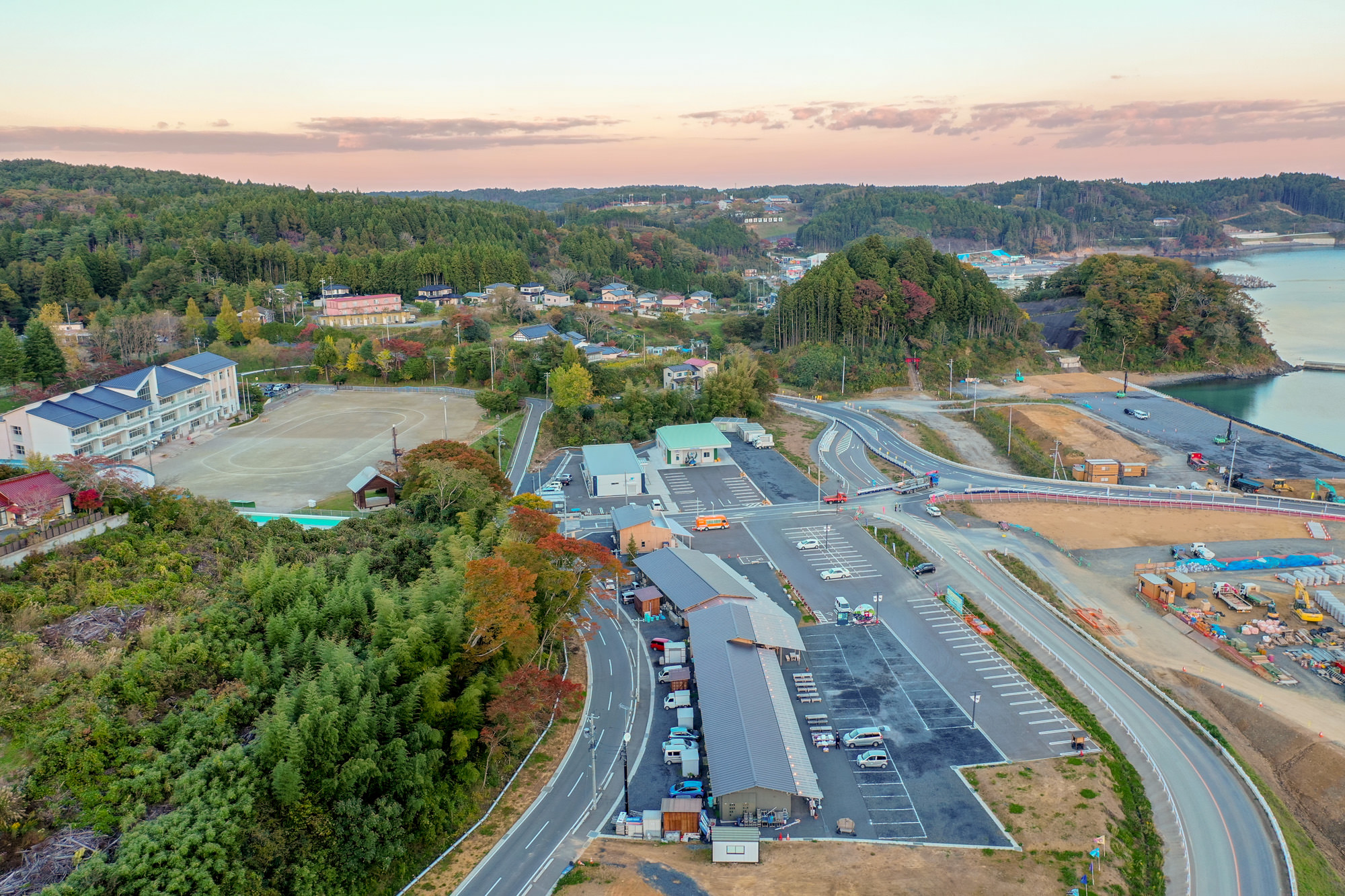 山々に囲まれた静かな町の風景で、中央には広い駐車場と複数の建物があり、その隣には運動場が広がっていて、自然に包まれた地域の様子が見える写真