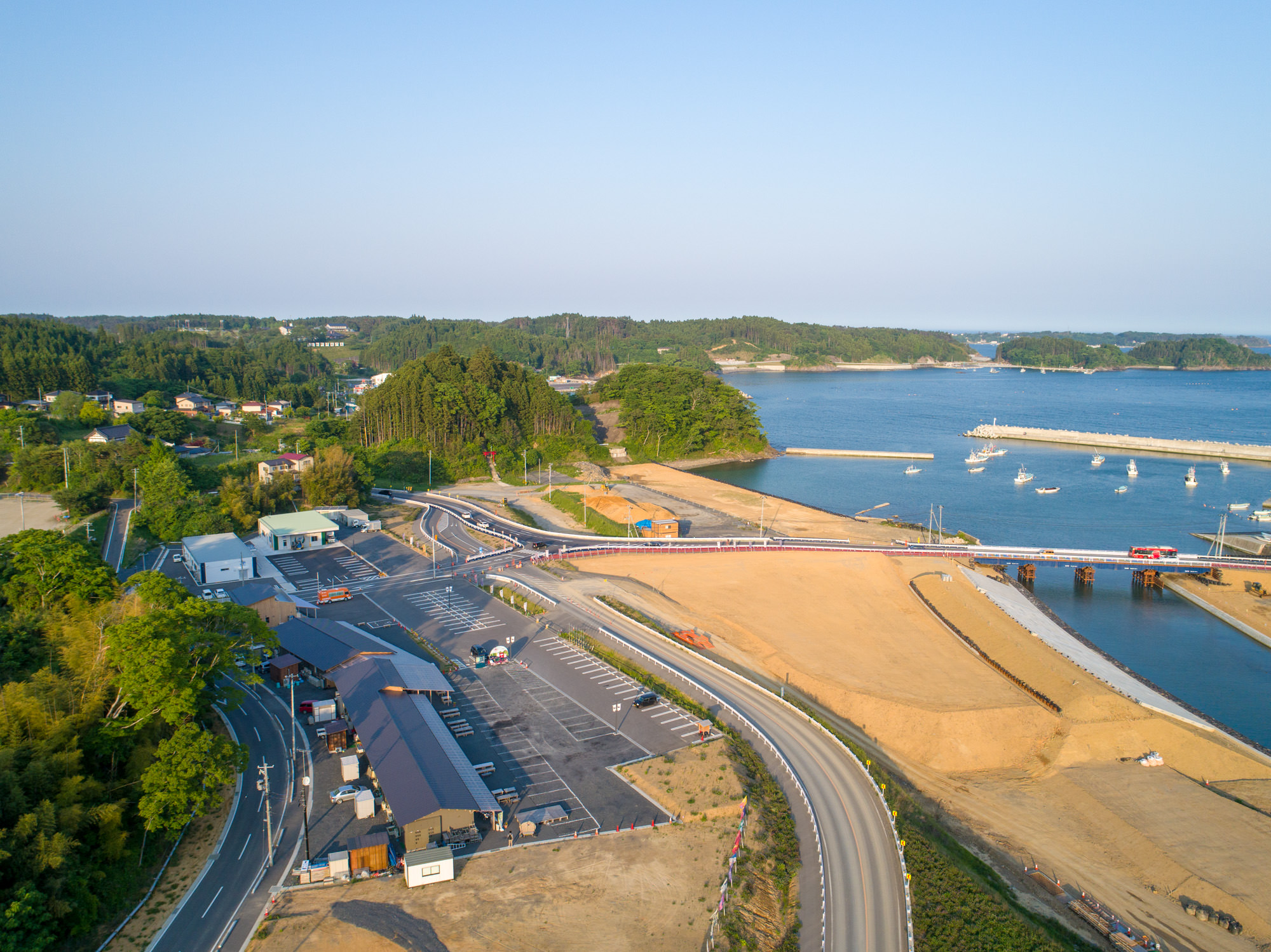 自然と人工構造物が混在する海沿いの地域を上空から撮影したもので、右側には桟橋に停泊する船と水域、橋が水上を渡って道路につながり、左側には建物や駐車場、道路が緑に囲まれて広がり、背景には森林が地平線まで続く風景の写真