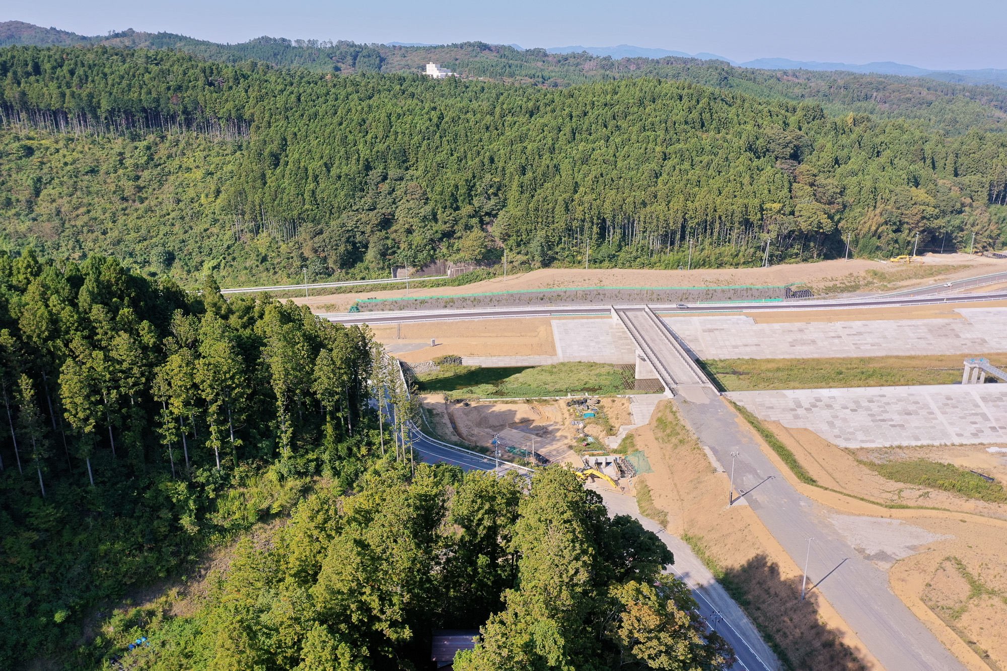山の中腹に切り開かれた場所で、橋と道路の建設が進んでいる様子を森と一体で映し出した、防災インフラ整備の途中経過の写真