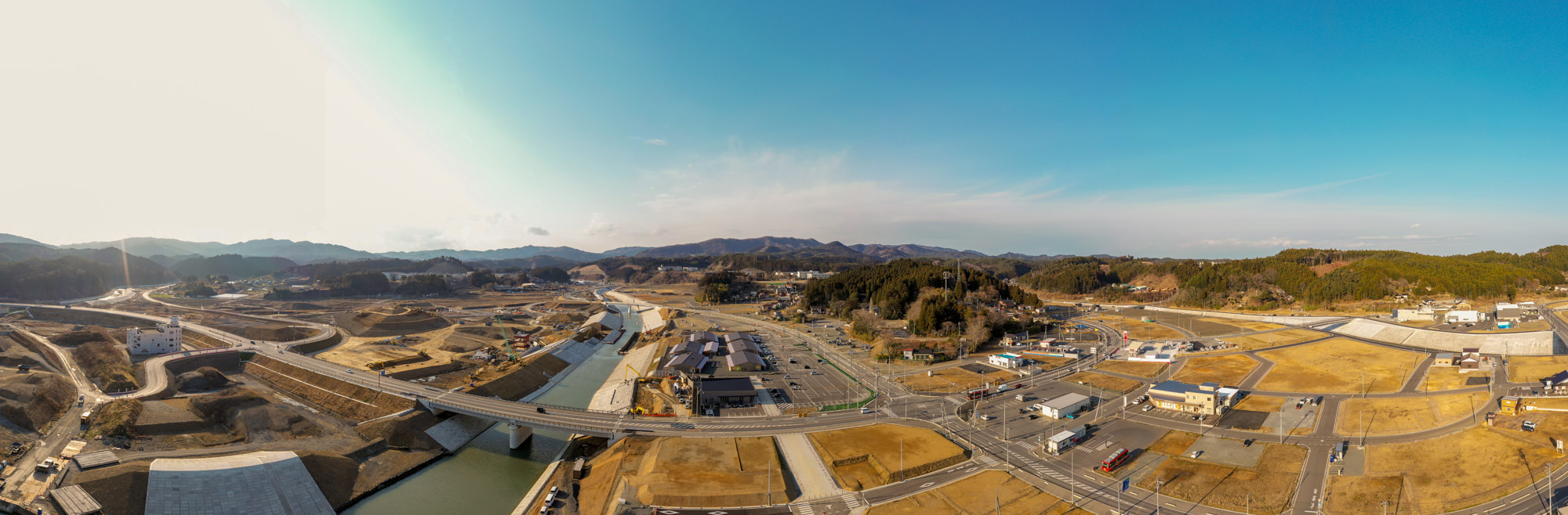 中央を流れる川に複数の橋が架かり、周囲には建物や構造物が点在し、遠くには山々が広がり、道路が地域を結ぶ晴天の農村地帯を上空から捉えた地方の河川地域の写真
