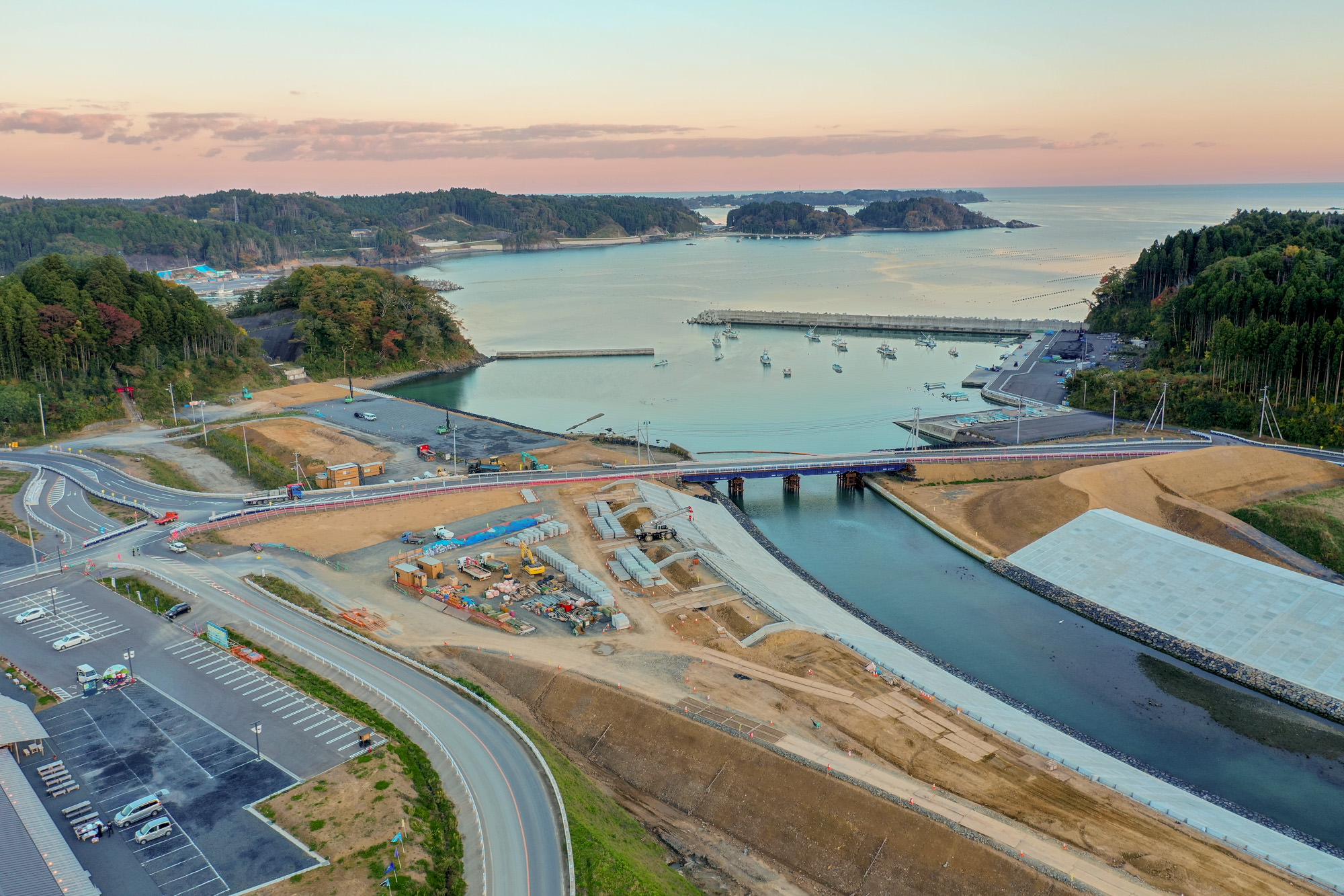 自然に囲まれた海沿いの風景で、川が海へと流れ込み、その上には橋が架かっており、周囲には工事現場や駐車場、道路が広がり、背景には木々に覆われた丘と水平線まで続く海が見え、空には少し雲が浮かぶ写真