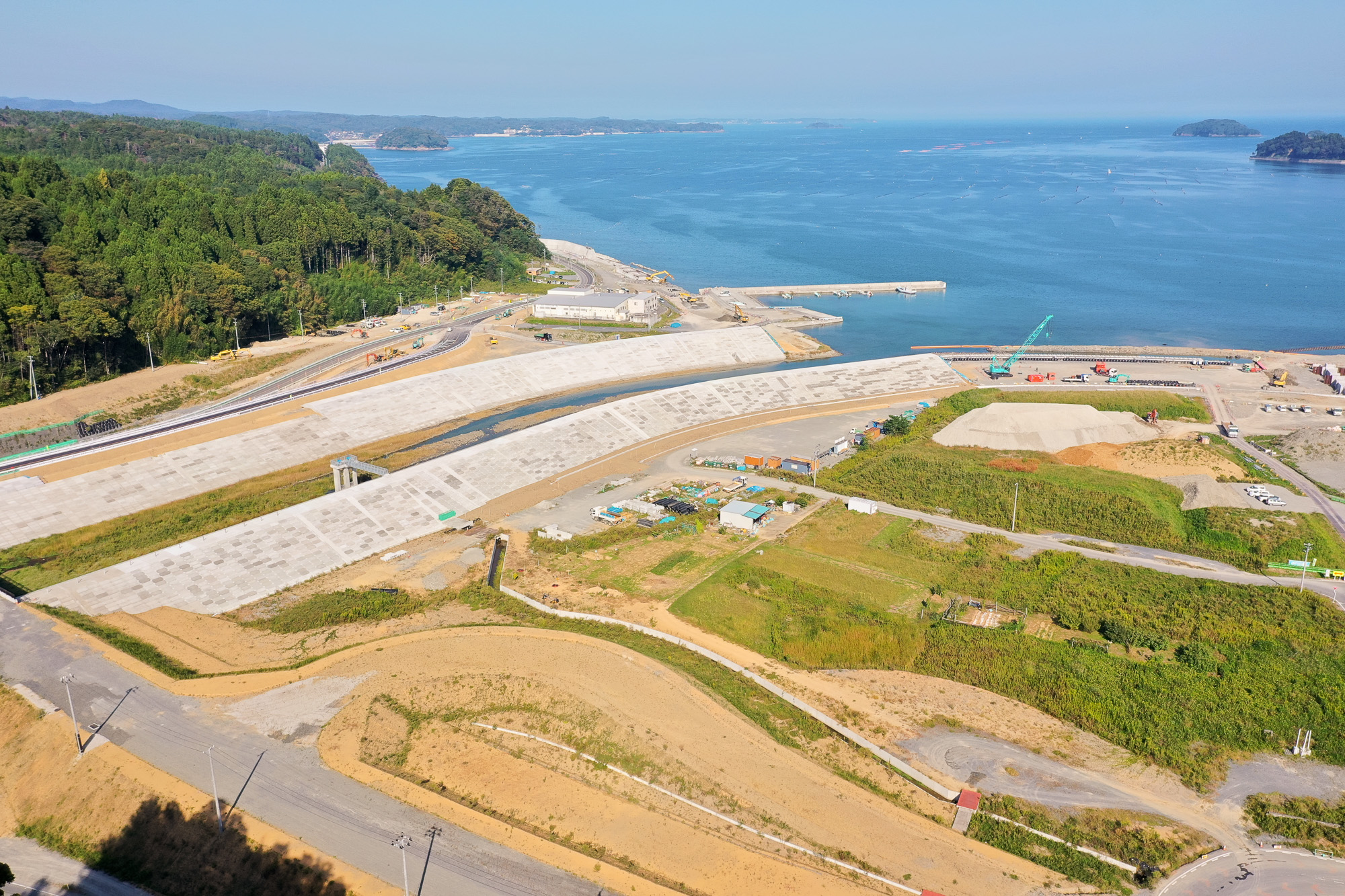 山に囲まれた湾の奥にある港で、防潮堤と工事中の土地整備、クレーンなどの重機が並ぶ工事現場を上空から見た海辺のインフラ再建の写真