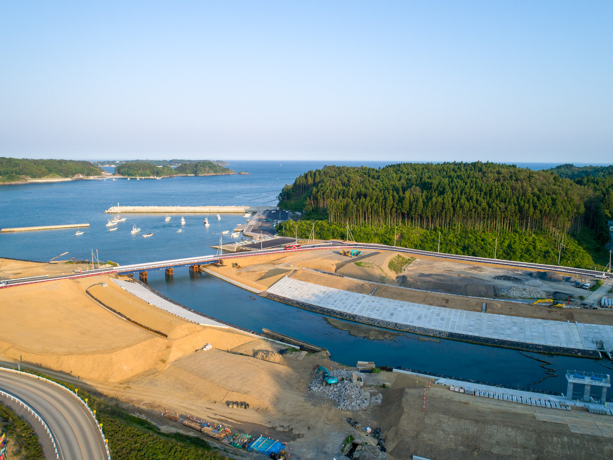 水上に架かる橋を中心とした海沿いの地域を上空から撮影したもので、建設資材や機材が並ぶ工事現場があり、周囲には森林と空き地、背景には桟橋や防波堤に停泊する複数の船と広がる海、晴れた空が見える風景の写真