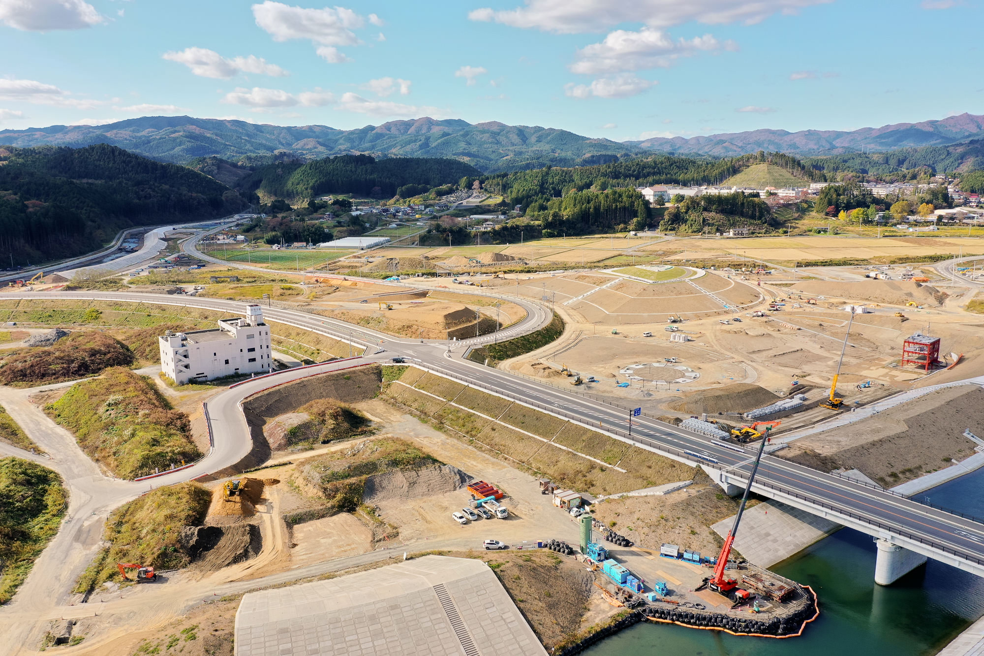 山々に囲まれた地域で、複数の道路が交差する造成中の広大な土地と、橋のたもとで作業するクレーン車や重機が見える復興工事現場の写真