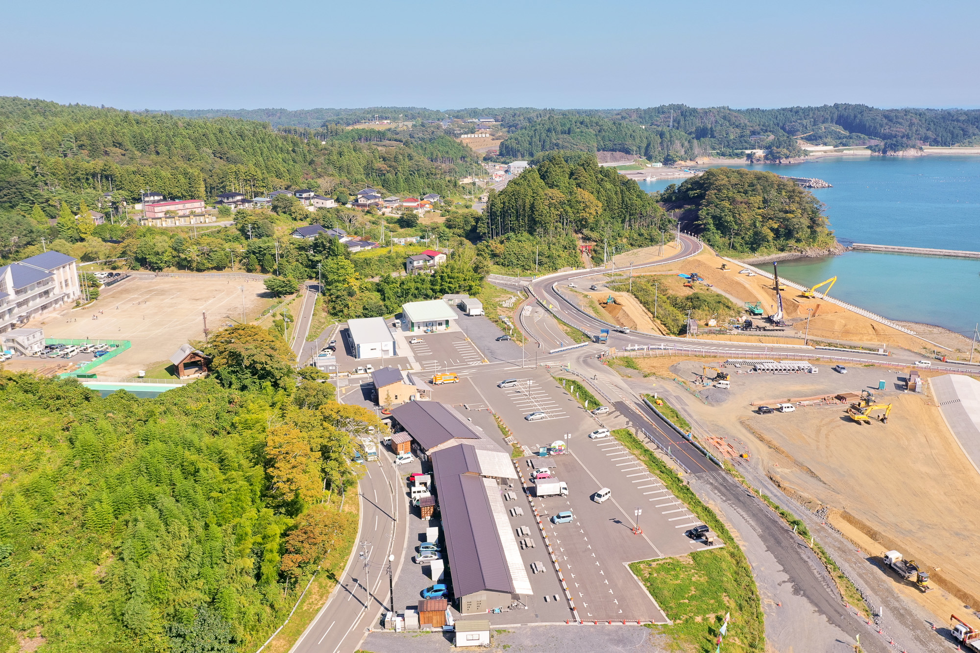 山のふもとの町並みと、グラウンドで遊ぶ子どもたちが見える学校、駐車場、曲がりくねった道路が見渡せる写