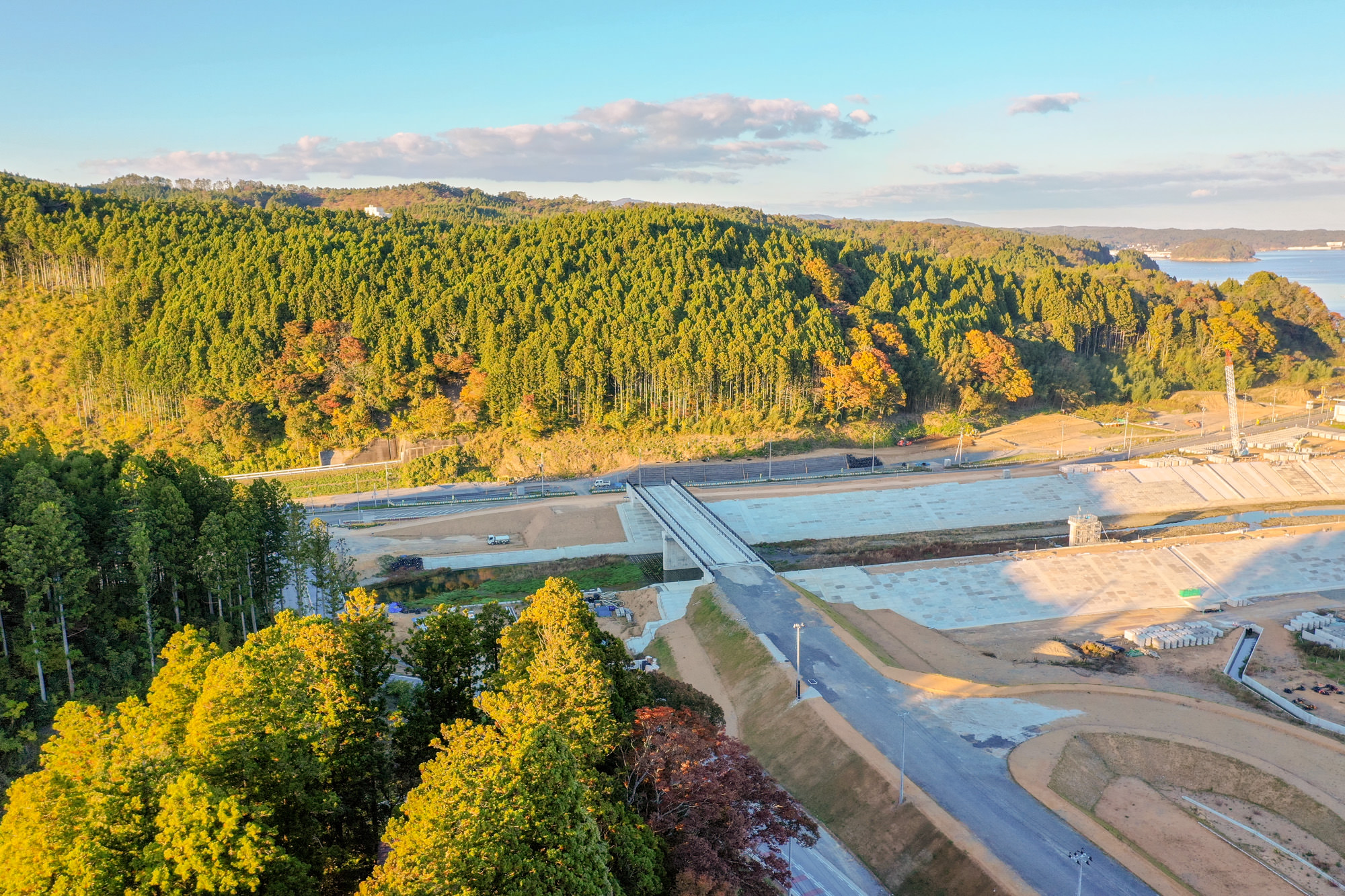 緑が多い山林の手前にコンクリートで整備された川と橋があり、周辺に整地された土地や道路が広がる風景の写真