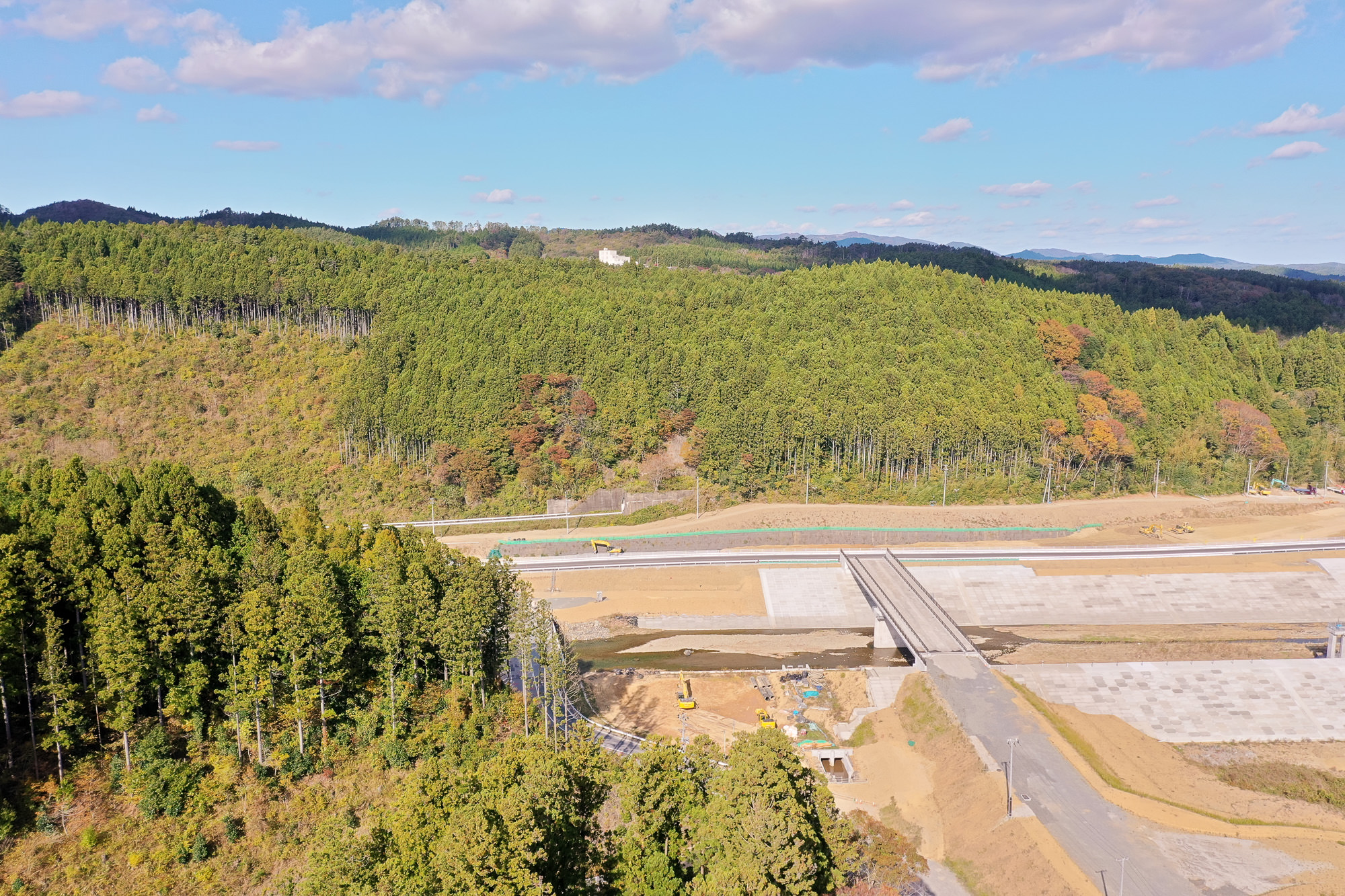 小さな川にコンクリートで整備された護岸と橋があり、周囲を緑の山々が囲み、手前には工事用重機が並ぶ現場の写真