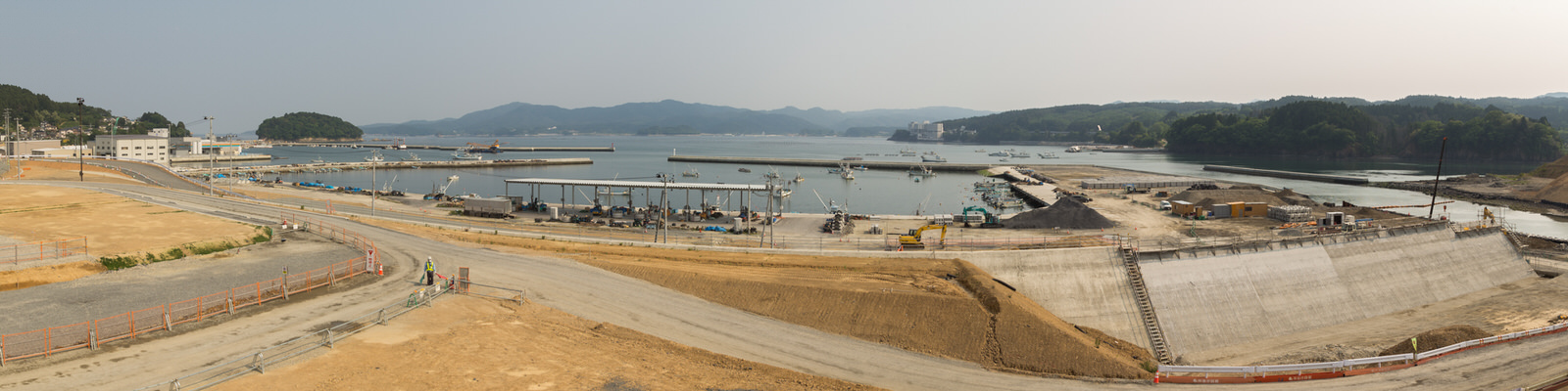 港に面した工事現場で、クレーンやトラック、土の山などの建設資材が並び、三方を水に囲まれた場所に複数の桟橋が伸び、船が停泊しており、背景には木々に覆われた丘が広がり、手前には大きなコンクリート構造物が建設中の様子のパノラマ写真