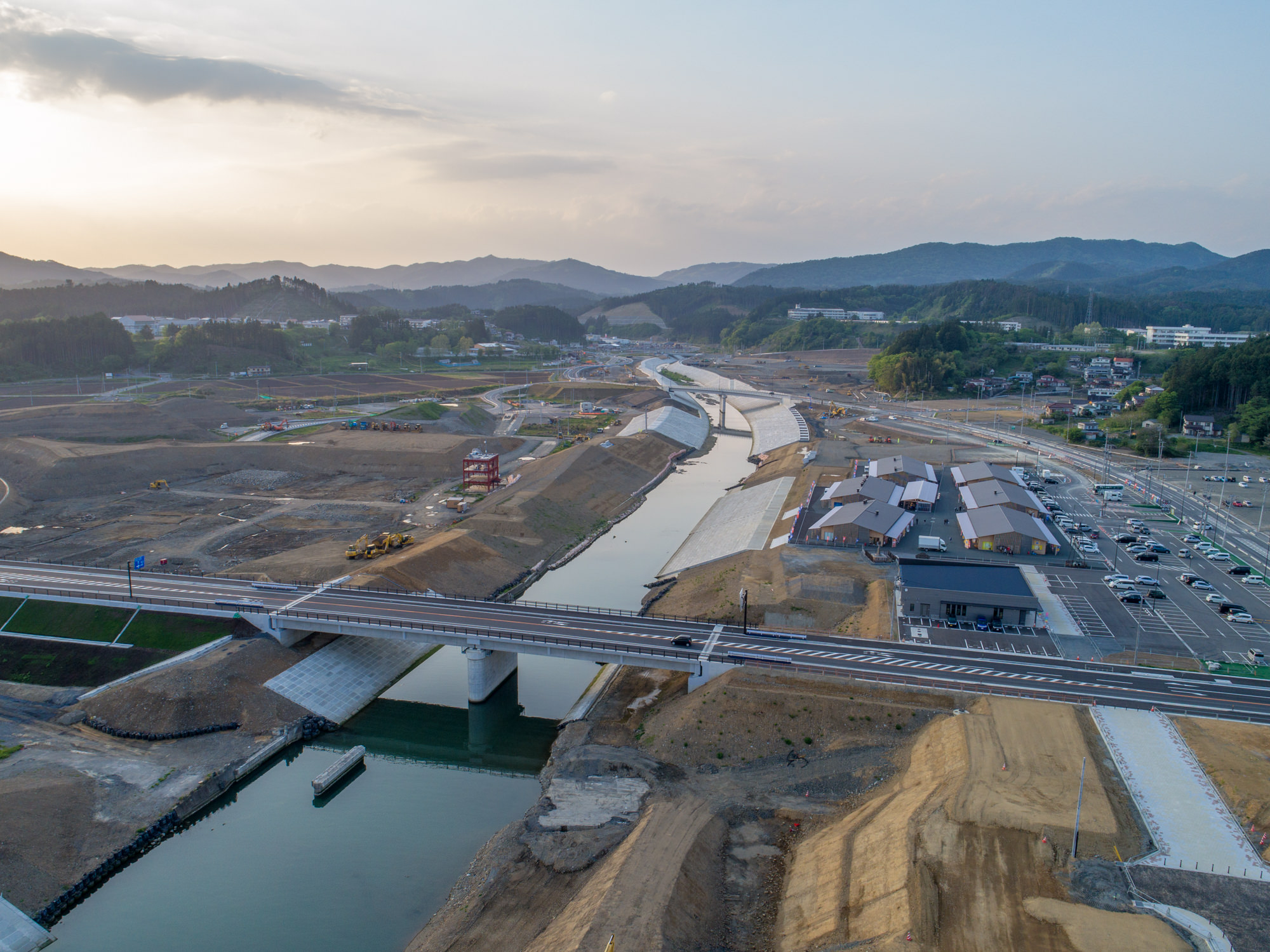 川が中央を流れる建設現場を上空から捉えた写真で、川には二本の橋が架かり、右側には複数の建物や駐車場が広がり、周囲には丘や森林、さらに奥には山々が連なり、空には雲が浮かび、朝または夕方の柔らかな光に照らされた風景の写真