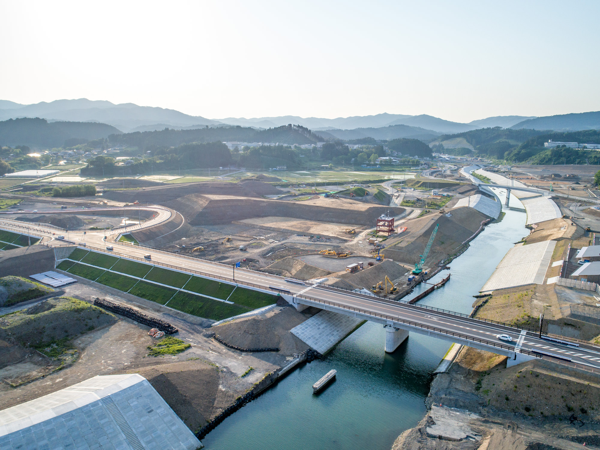 川に架かる橋を中心とした建設現場を上空から撮影したもので、周囲には建設機械や未完成の構造物、土工が広がり、背景にはなだらかな丘と山々が見える風景の写真