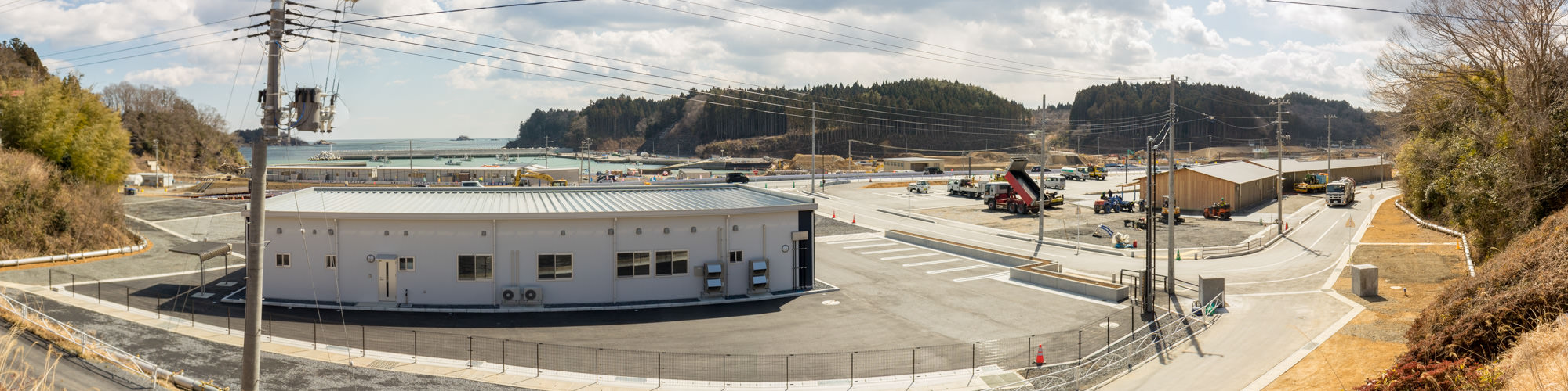 中央には白い平屋の建物があり、その周囲には舗装された道路や駐車スペースが広がって、背景には海と緑豊かな丘陵地帯が広がる海に近い建設現場を写した写真