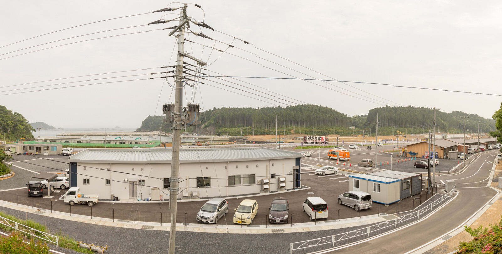 曇り空の下、複数の車が停まる駐車場の奥に平屋の建物があり、高い電柱から複数の電線が伸び、背景にはさらに建物や車両、森林が広がる、郊外の施設周辺を上空から捉えた写真