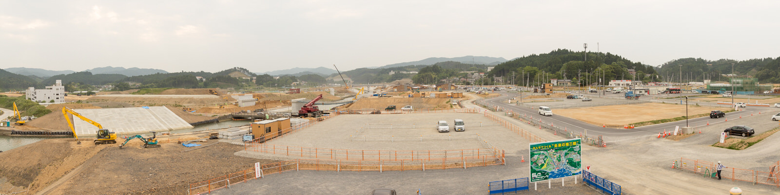 丘と木々に囲まれた地域で、クレーンやショベルカーなどの建設機材が点在する広大な工事現場を上空から捉え、手前には車が停められた柵で囲まれたエリアと地図が描かれた大きな看板が見える、開発が進む地域のパノラマ写真