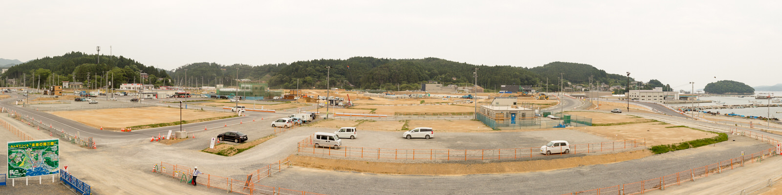 曇り空の下、海沿いの工事現場で白いバンや車が複数停まっており、仮設フェンスに囲まれた敷地内には建設資材や機材が点在し、左側には工事内容を示す看板が設置され、背景には木々に覆われた丘と建物が広がる、開発中の沿岸地域を上空から捉えたパノラマ写真