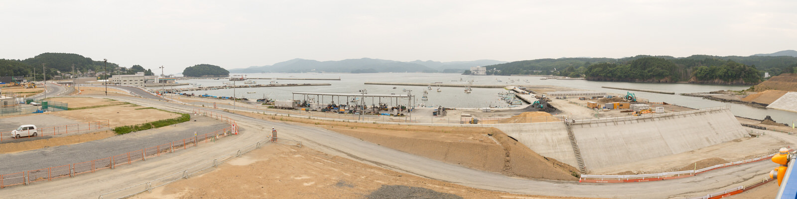 曇り空の下、緑に覆われた丘を背景に、海岸沿いで複数の小型船が停泊する水辺に面した工事現場が広がり、コンクリート構造物や道路の建設・補修が進められている様子を上空から捉えた、沿岸開発の風景のパノラマ写真