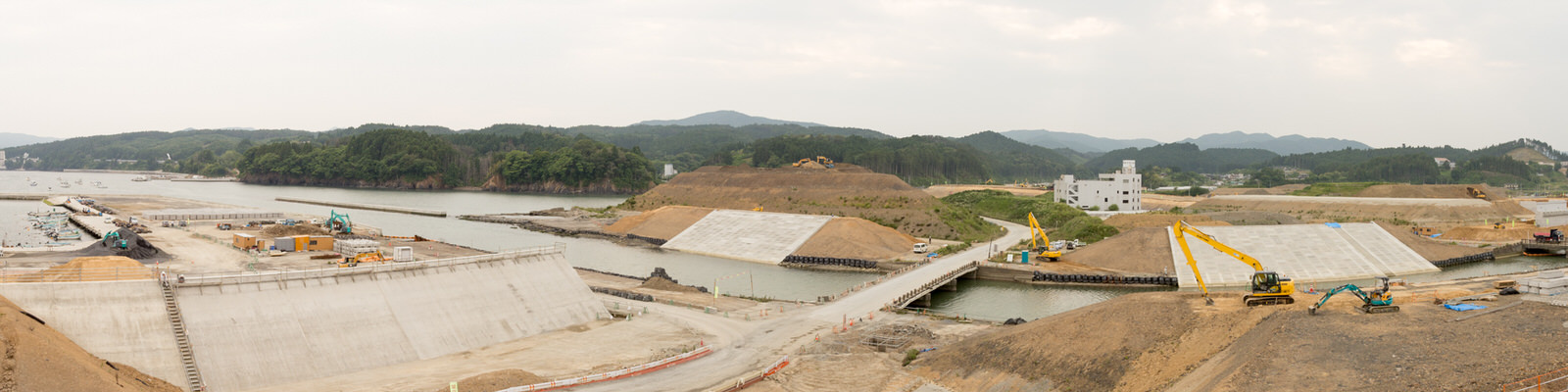 曇り空の下、川や運河沿いの工事現場で、傾斜した壁や平らな基礎などの大きなコンクリート構造物が並び、ショベルカーやクレーンなどの建設機材が稼働し、背景には森林に覆われた丘や山が広がる、水辺のインフラ整備が進む様子を上空から捉えたパノラマ写真