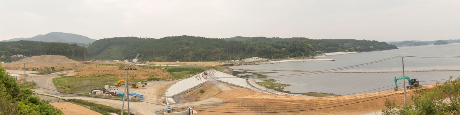 右側に海岸線が広がる沿岸地域で、クレーンやショベルカーなどの建設機材が点在し、整地された土地や建設途中の構造物、自然の木々や水辺が混在し、背景には丘と森林が広がり、電線も横切る、開発と自然が交差する風景を上空から捉えたパノラマ写真