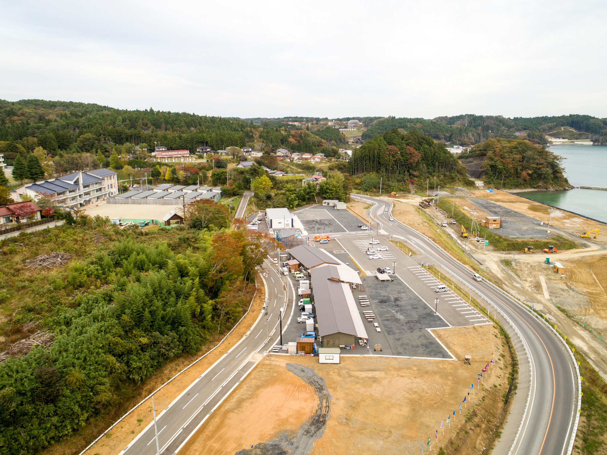 新しく整備された駐車場と建物が並ぶ小さな海沿いの町を上空から捉え、周囲には豊かな緑と丘が広がり、背景には海が見える写真