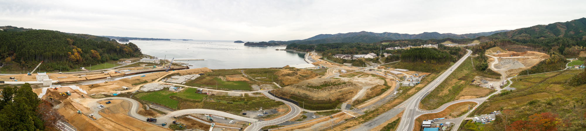 海沿いの建設現場を広角で捉えたパノラマ写真で、道路や建物、緑地が点在し、背景には森に覆われた丘と山々、さらに水辺が広がる曇り空の下の写真