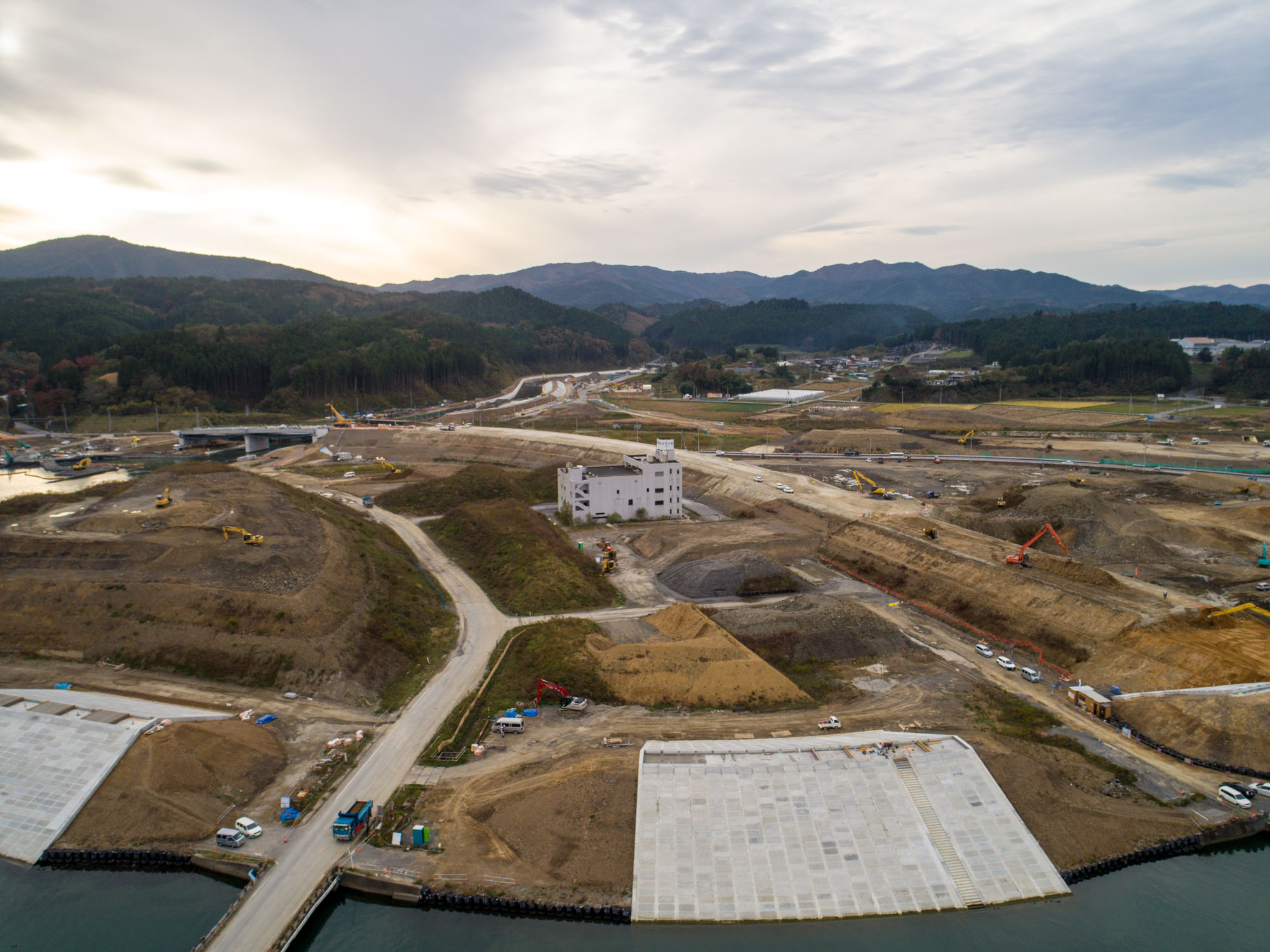 中央の建物を囲むように道路や重機、土木作業が広がる大規模な建設現場を谷間に位置する風景として上空から捉え、背景には山々が広がる曇り空の下の写真