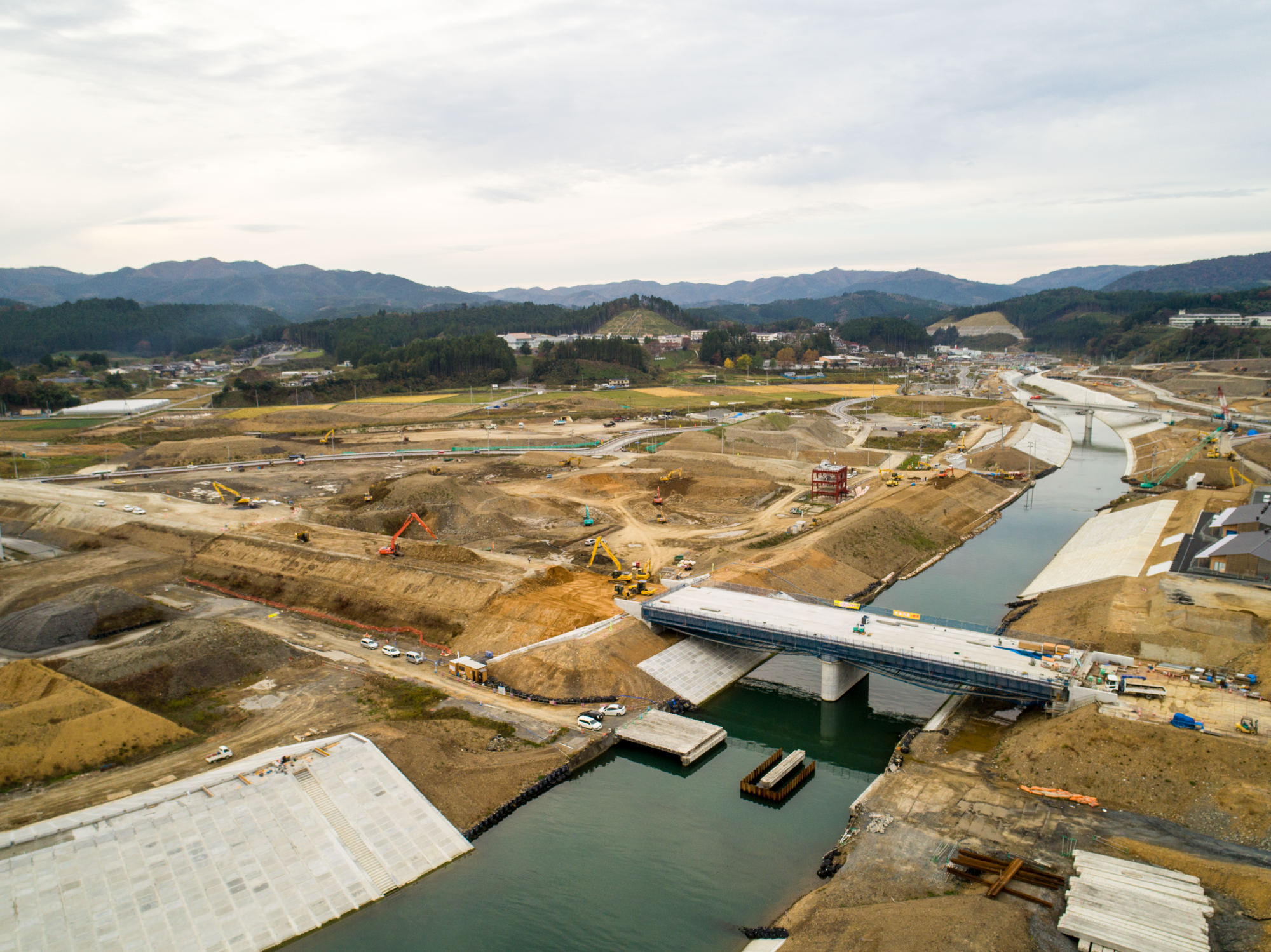 川に架かる建設中の橋と周囲の重機や資材が広がり、背景には丘や山々、整地された土地と緑が混在する曇り空の下の建設現場の写真