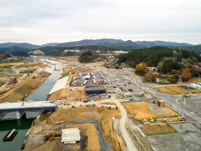 川を横切るように建設中の橋や、複数の車が駐車している駐車場が完備された商業施設の傍で沢山の重機が稼働している川沿いに広がる大規模な建設現場と、その周囲の自然風景を上空から捉え、遠くには連なる山々が見える写真