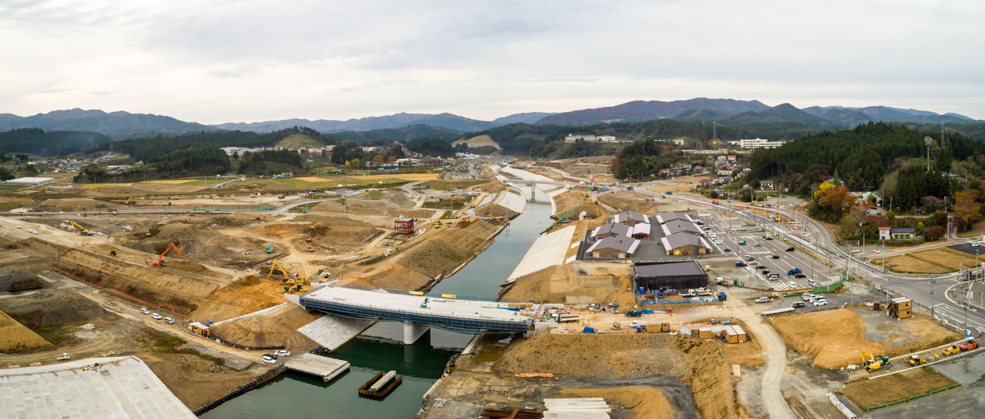 川に架かる橋の建設が進む大規模な工事現場を中心に、重機や資材、周囲の建物や道路、そして背景に広がる丘と山々が見えるパノラマ写真