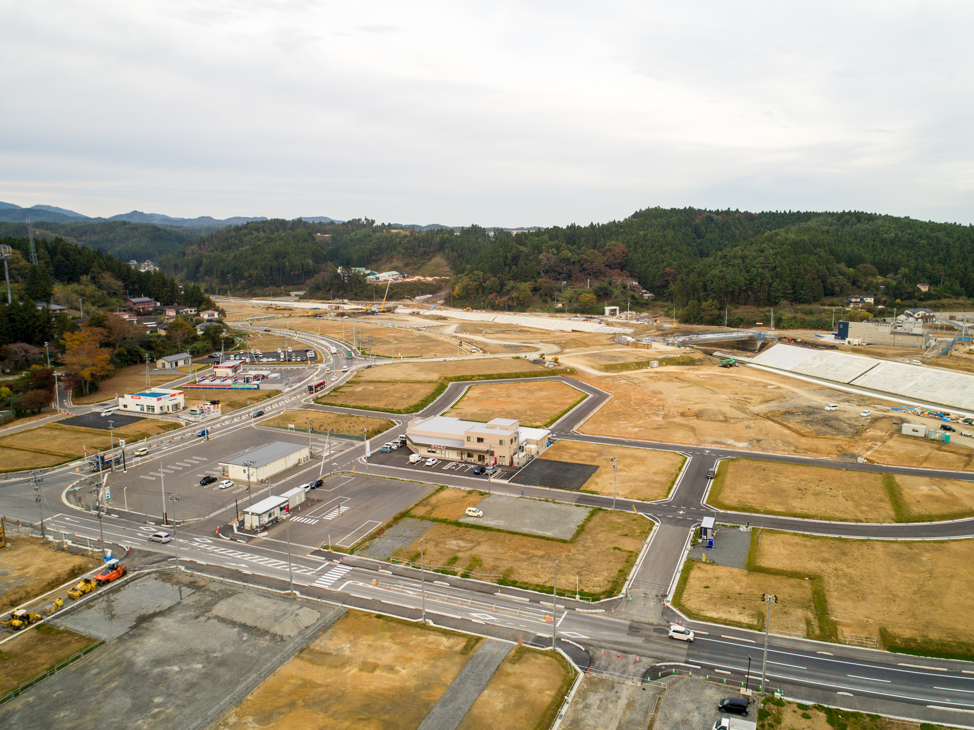 新しく整備された道路や空き地、点在する建物が広がる郊外の開発地域を上空から捉え、周囲には木々に覆われた丘と曇り空が広がる写真