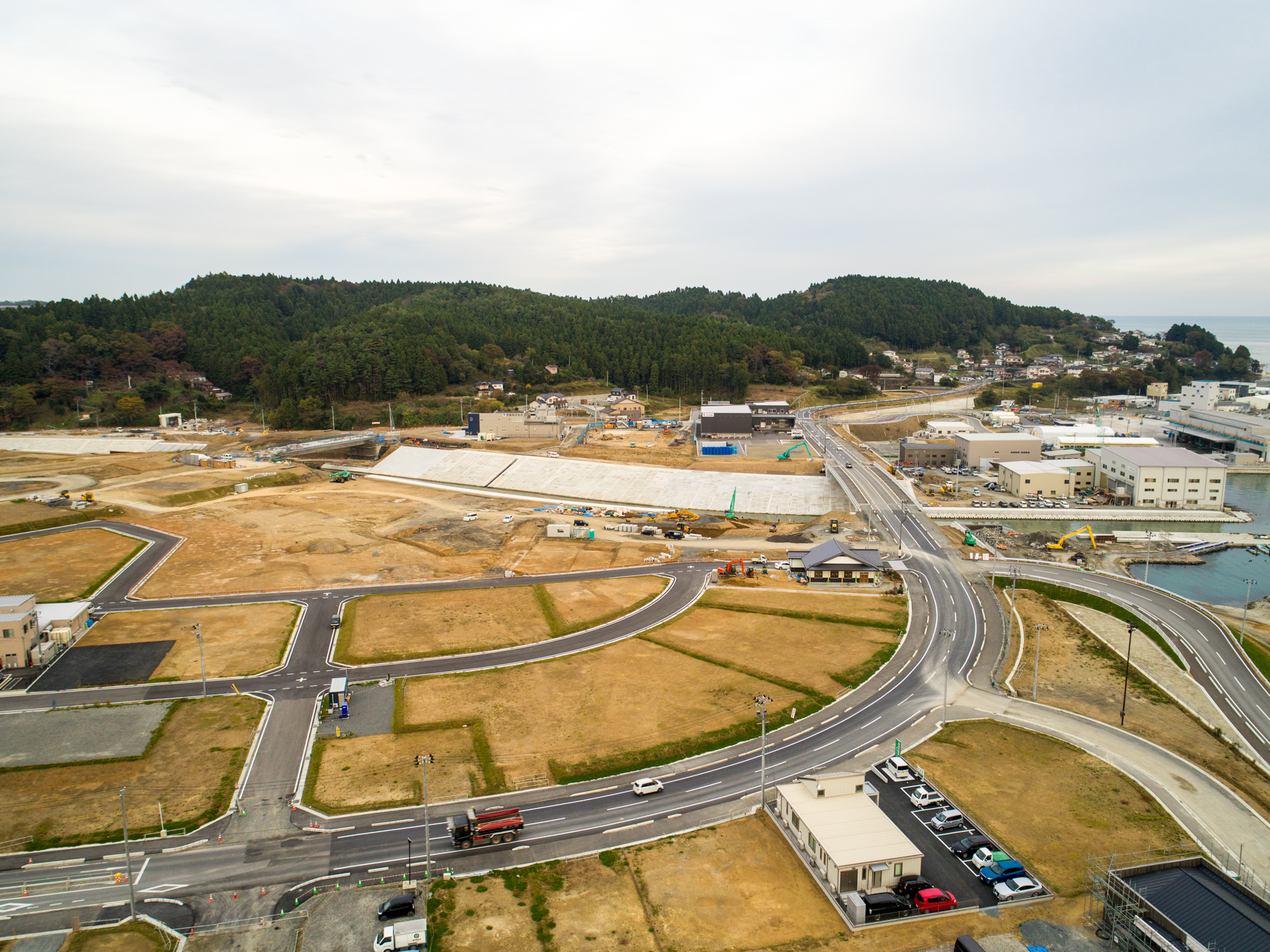 新しく整備された道路と区画が広がる海沿いの町を上空から捉え、森林に覆われた丘に囲まれ、建物や建設機械が点在し、水辺に架かる橋へと続く曲がりくねった道が印象的な写真