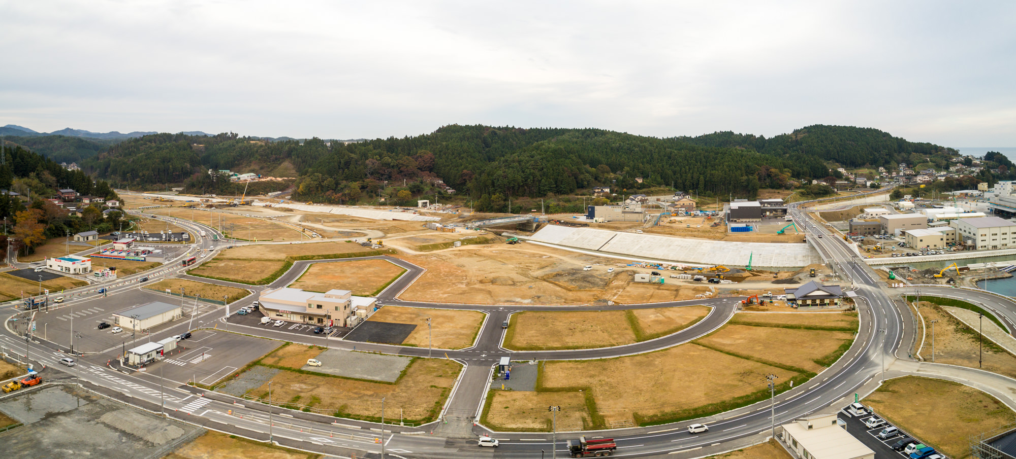 道路や建物、空き地が広がる志津川地区をパノラマで捉え、開発地と未開発地が混在し、森林の丘と建物が背景に広がる曇り空の下の写真