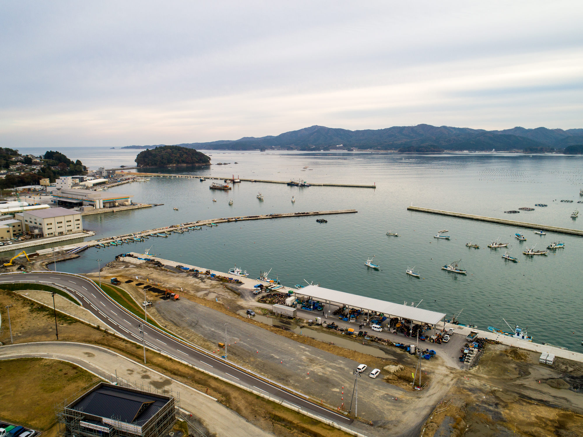 複数の船が浮かぶ海辺の港を中心に、工業的な建物と空き地が広がり、曲がりくねった道が港へと続き、背景には山と島が穏やかな海に浮かぶ曇り空の下の写真